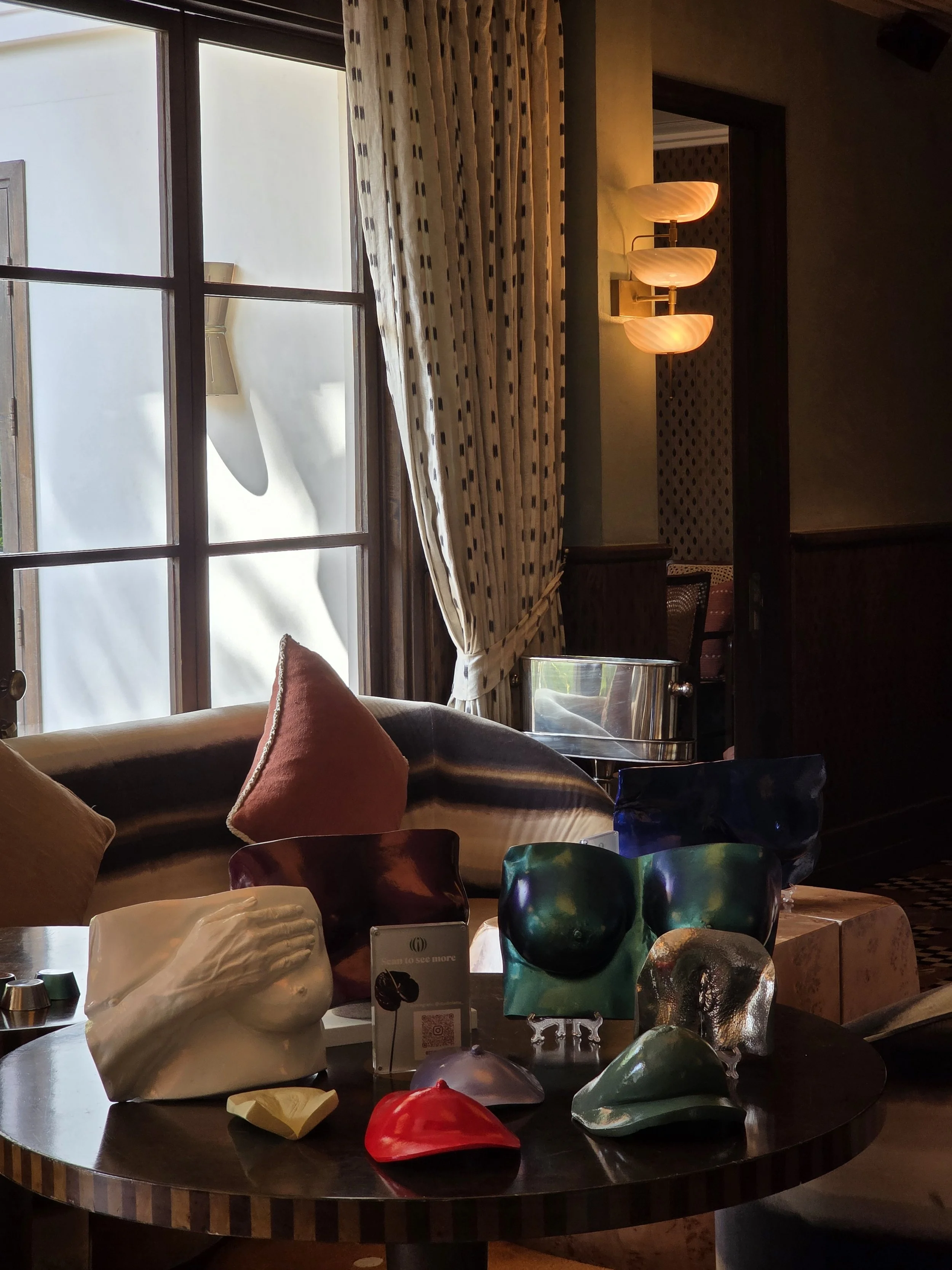 A collection of finished breast casts in various finishes — white plaster, iridescent teal, deep blue, and metallic — displayed on a round table with a QR code stand and Soho House interior in the background