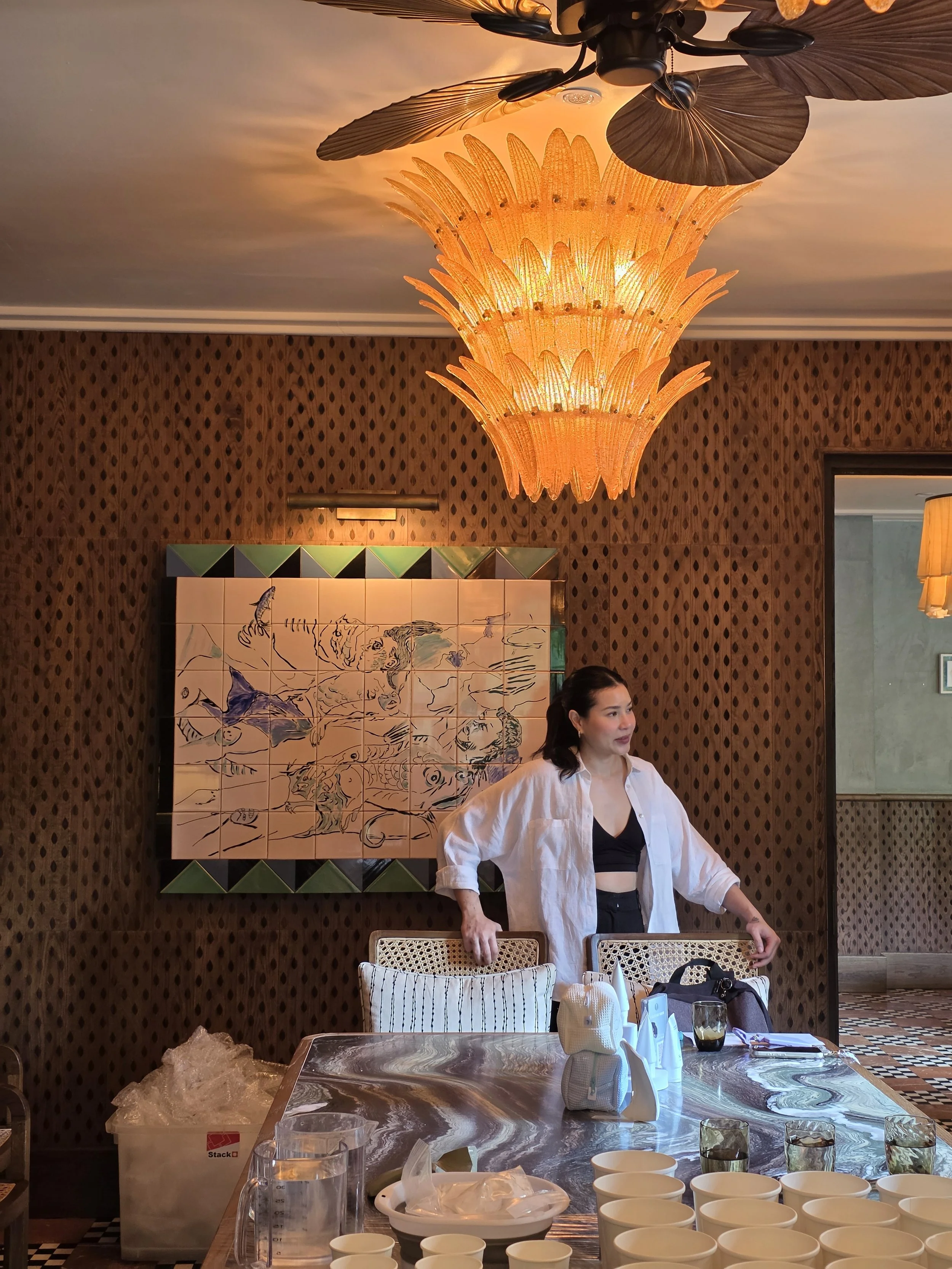 StudioLams_ artist standing at the marble casting table during setup at Soho House Bangkok, with white cups, plaster molds, and the ornate chandelier and tile artwork visible behind her