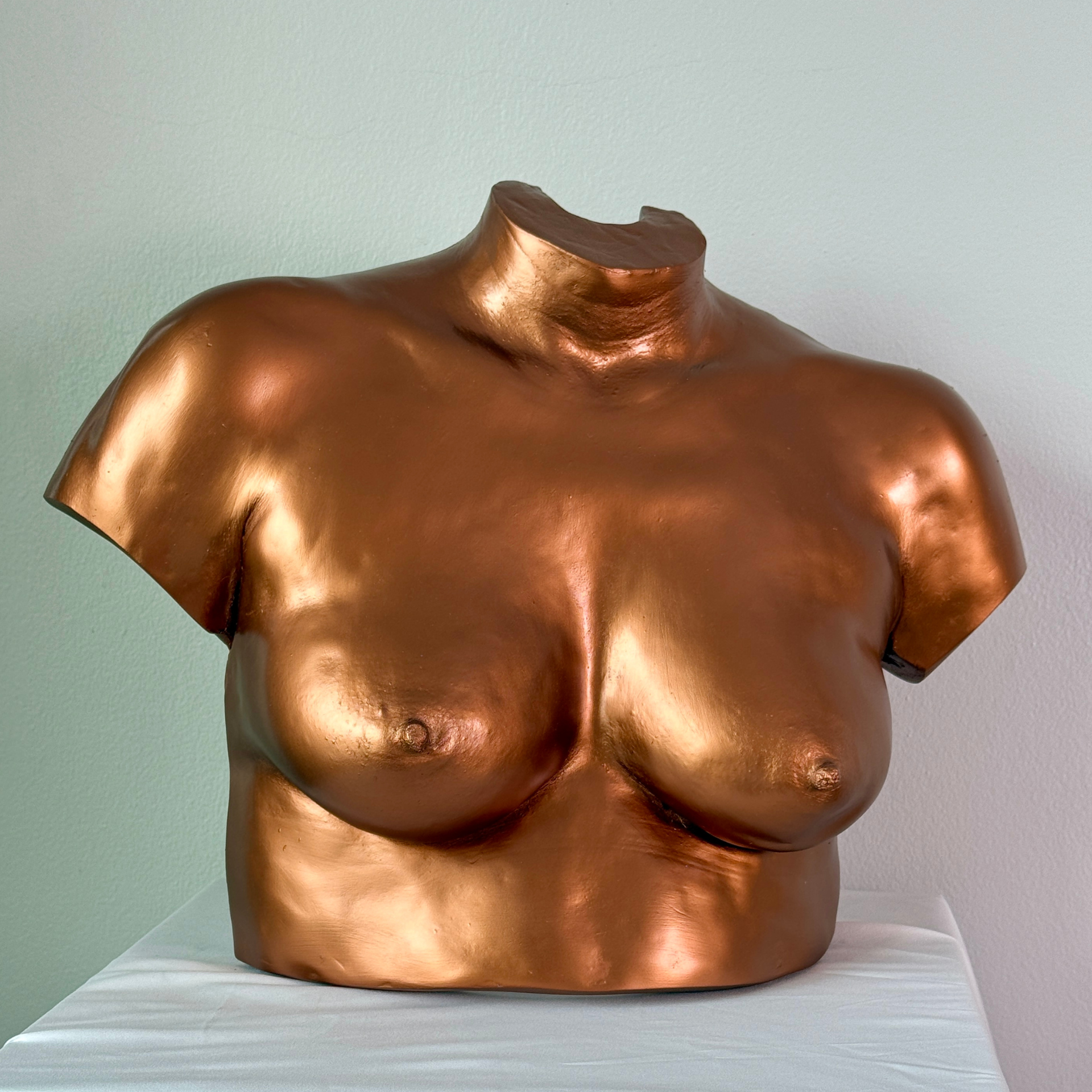 A sculpture of a woman's torso painted in metallic copper color, featuring detailed breasts and collarbone, placed on a white cloth surface against a plain light background.