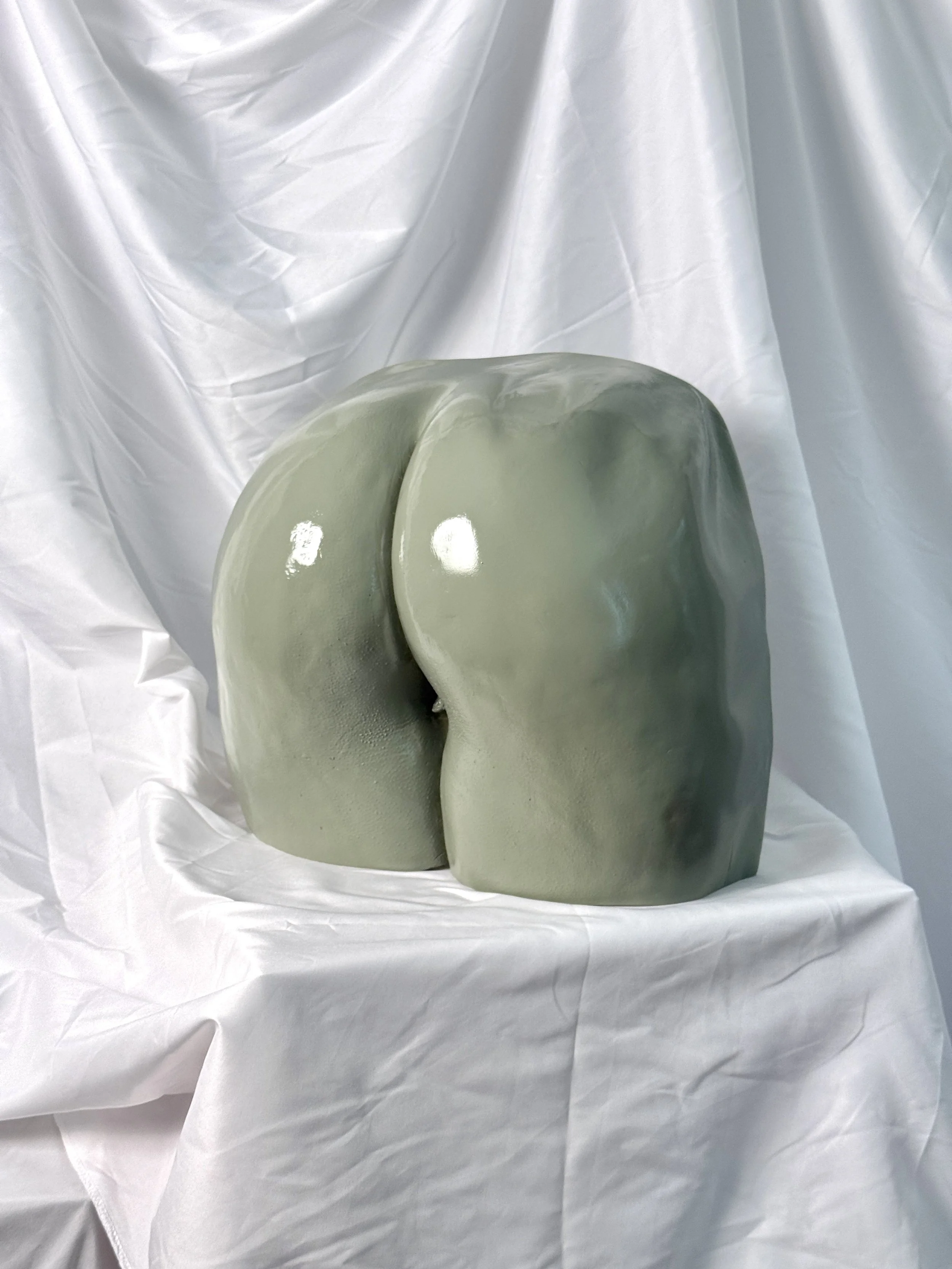 A shiny, greenish-gray bum casting sculpture, placed on white crumpled fabric.