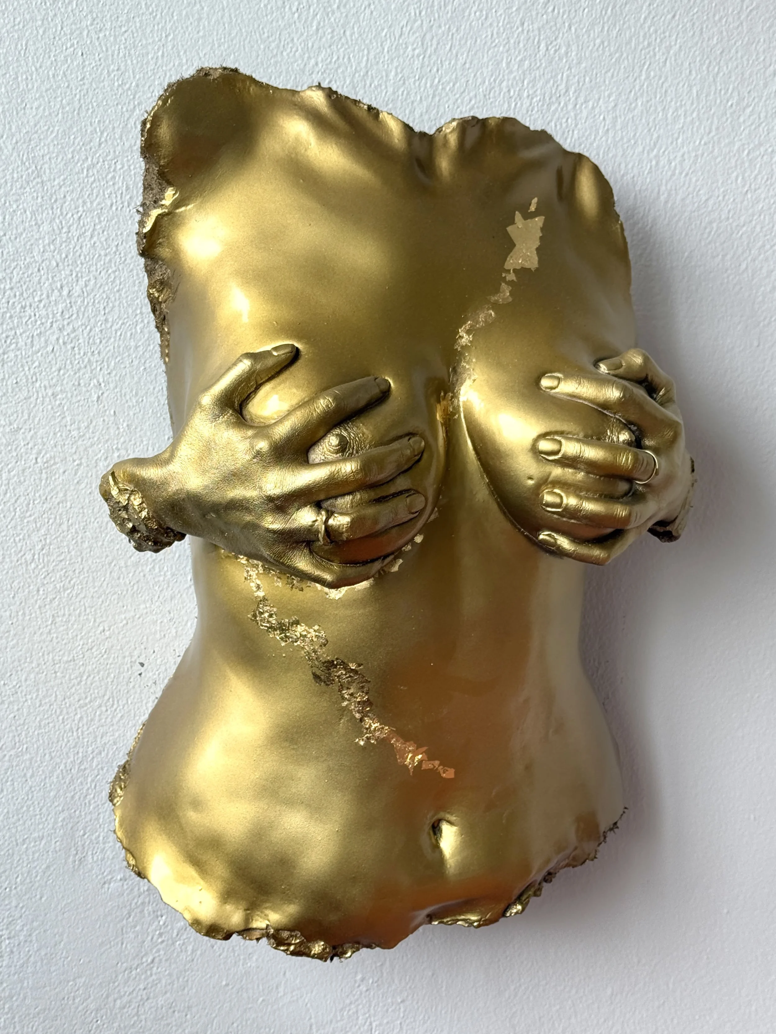 A sculpture of a human torso and head, painted gold, with hands covering the chest, mounted on a plain white background.