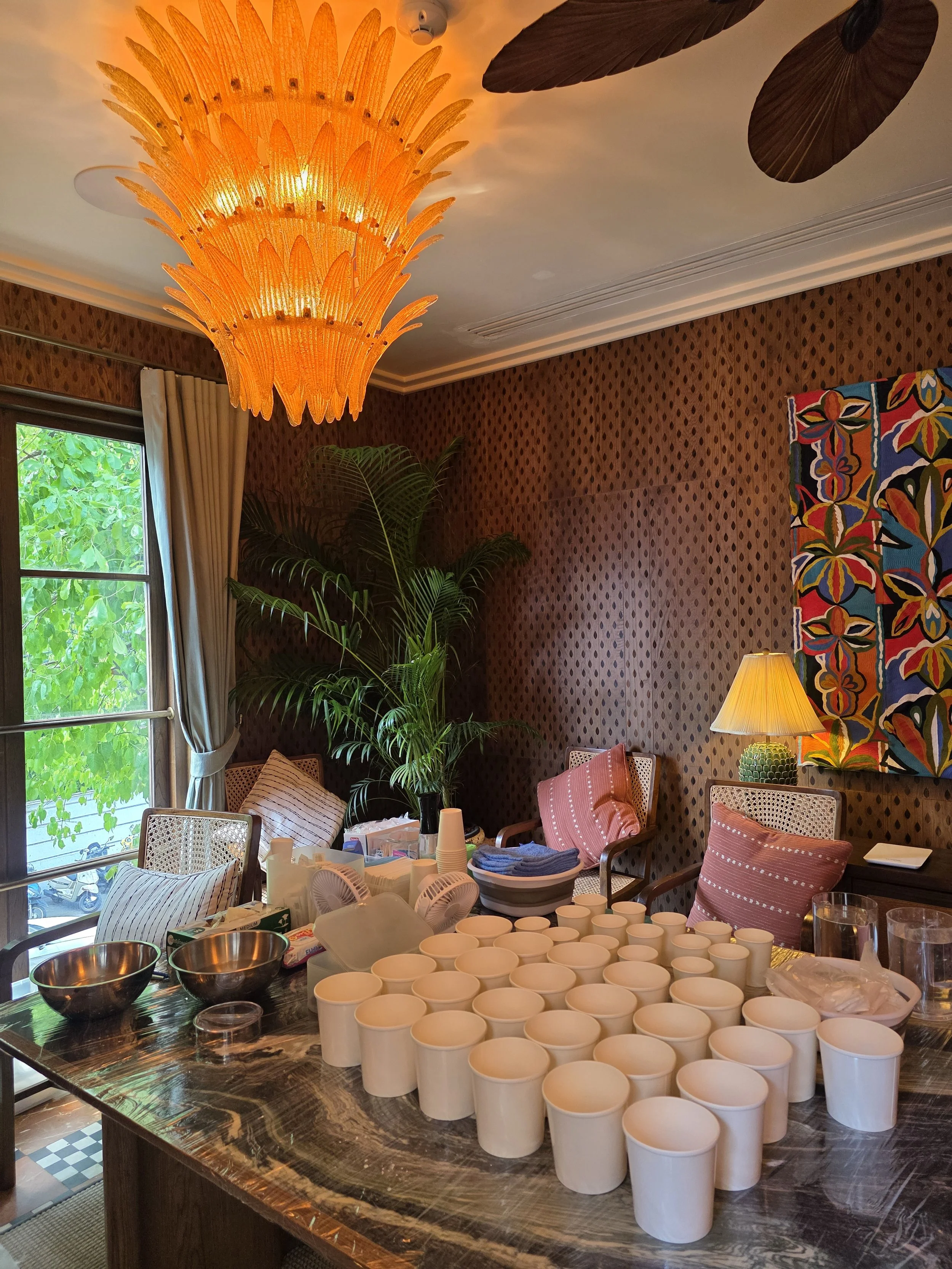 Wide view of the private casting room at Soho House Bangkok with rows of white cups ready on a marble table, rattan chairs, tropical plants, and a dramatic amber leaf chandelier overhead
