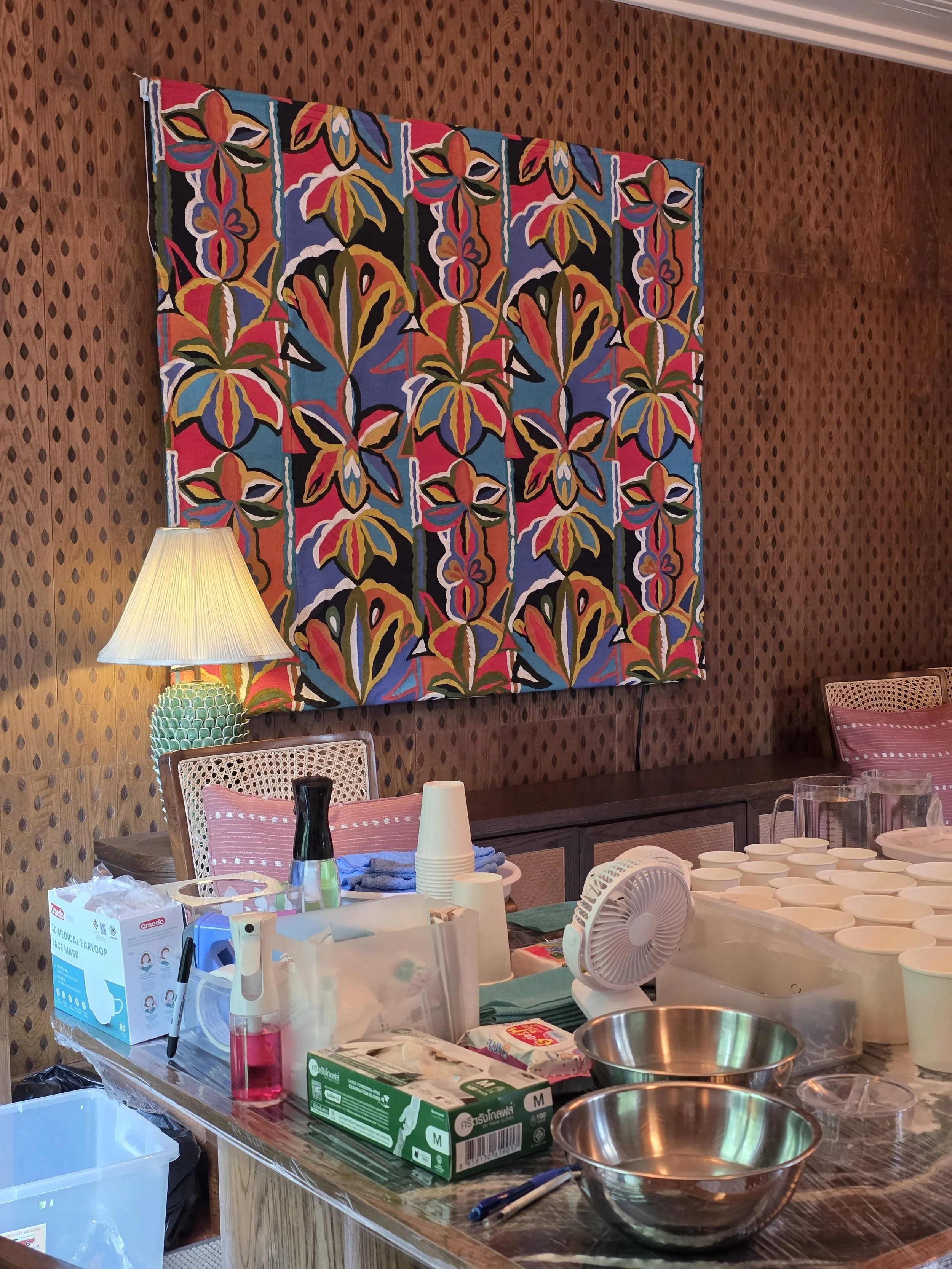 Casting supplies laid out on a marble counter at Soho House Bangkok, including mixing bowls, gloves, cups, and a spray bottle, with a colorful floral tapestry and warm lamp in the background