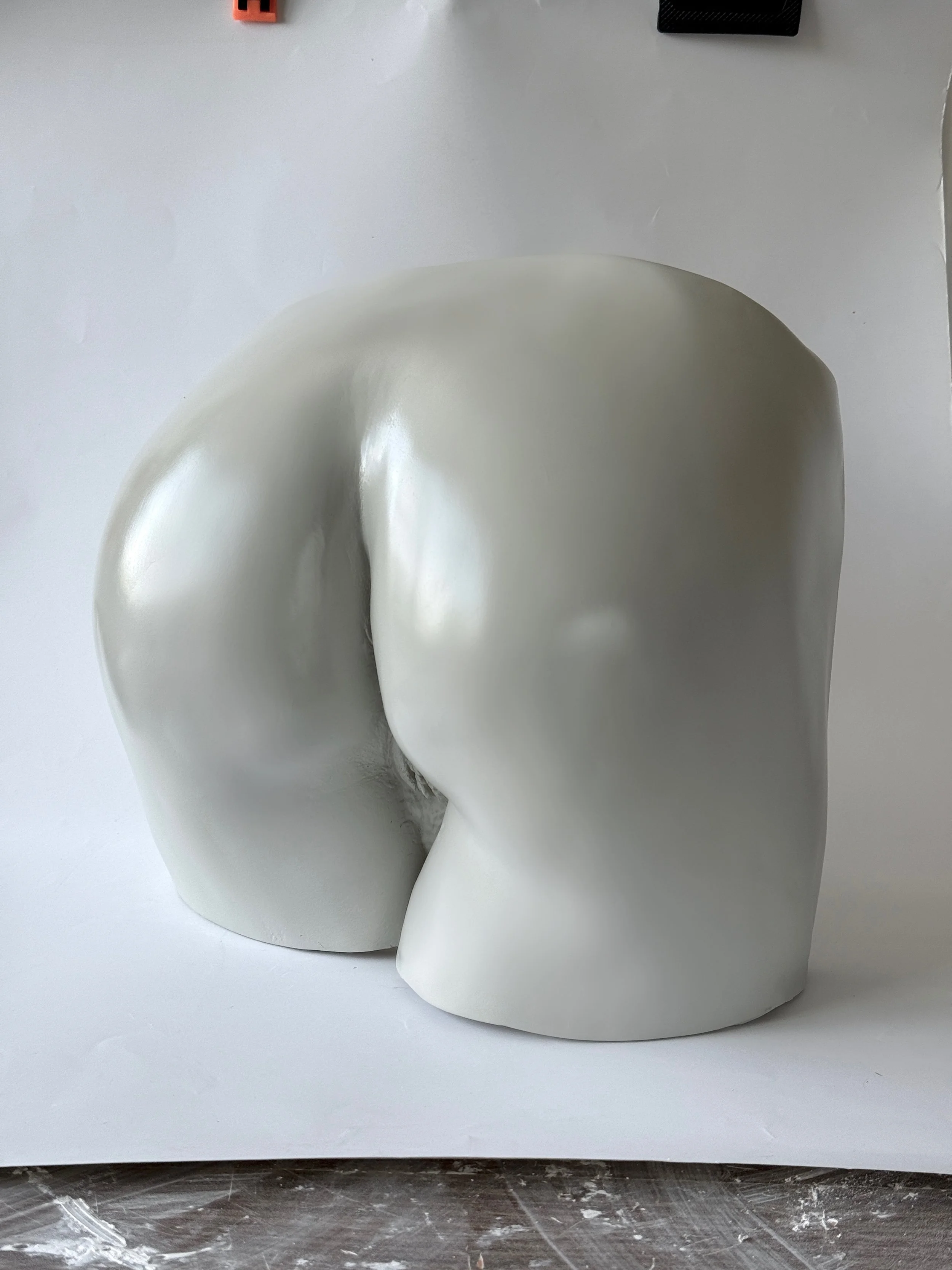 A white ceramic sculpture of a stylized human head without facial features, viewed from the side on a white surface, with a black and an orange object partially visible at the top.