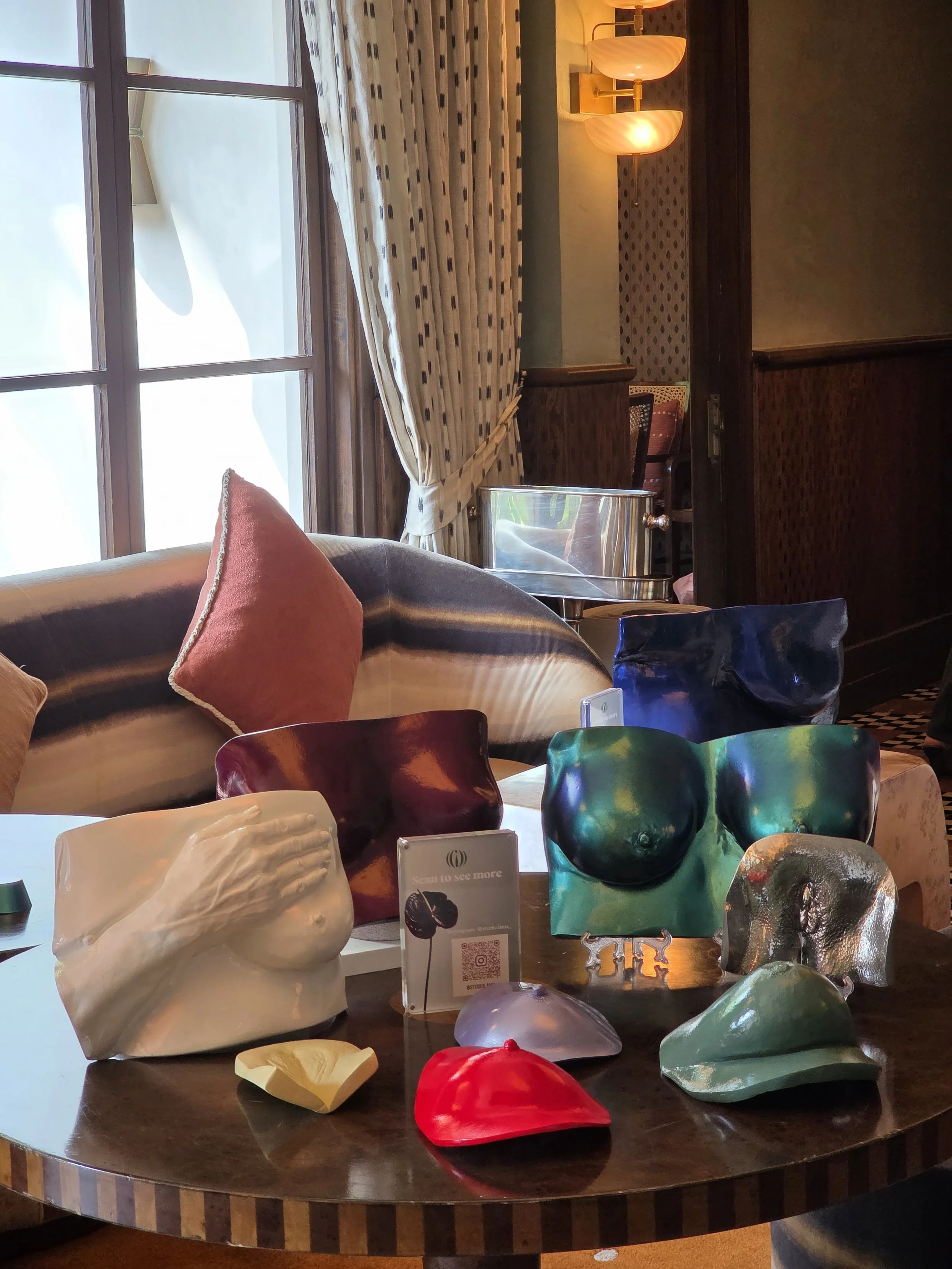 Display of colorful glass sculptures and ceramic art pieces on a round wooden table in a gallery setting, with a large window, curtains, and warm lighting in the background.