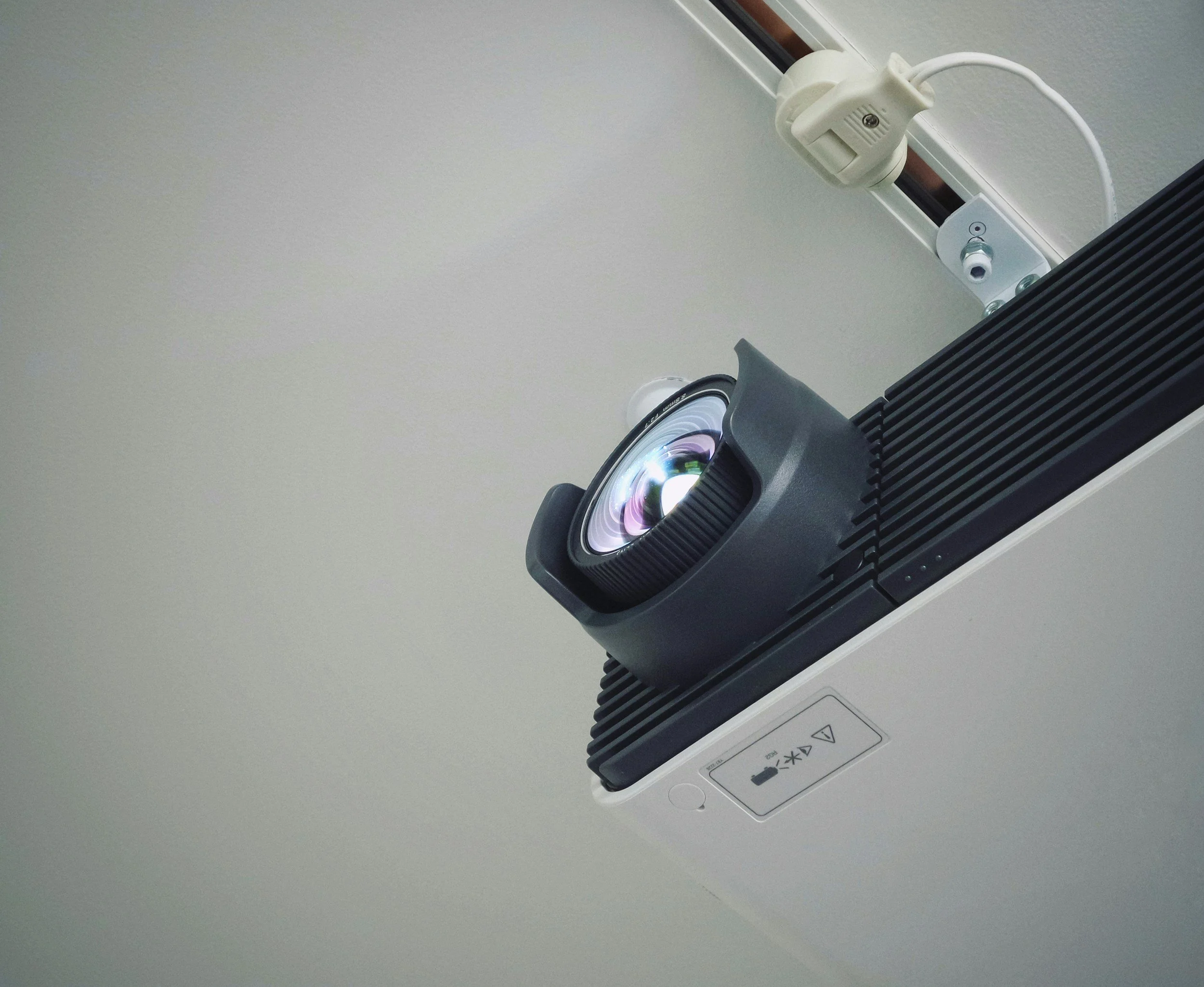 Close-up of a ceiling-mounted projector with a lens emitting light, connected to a power strip with a plug, mounted on a white ceiling.