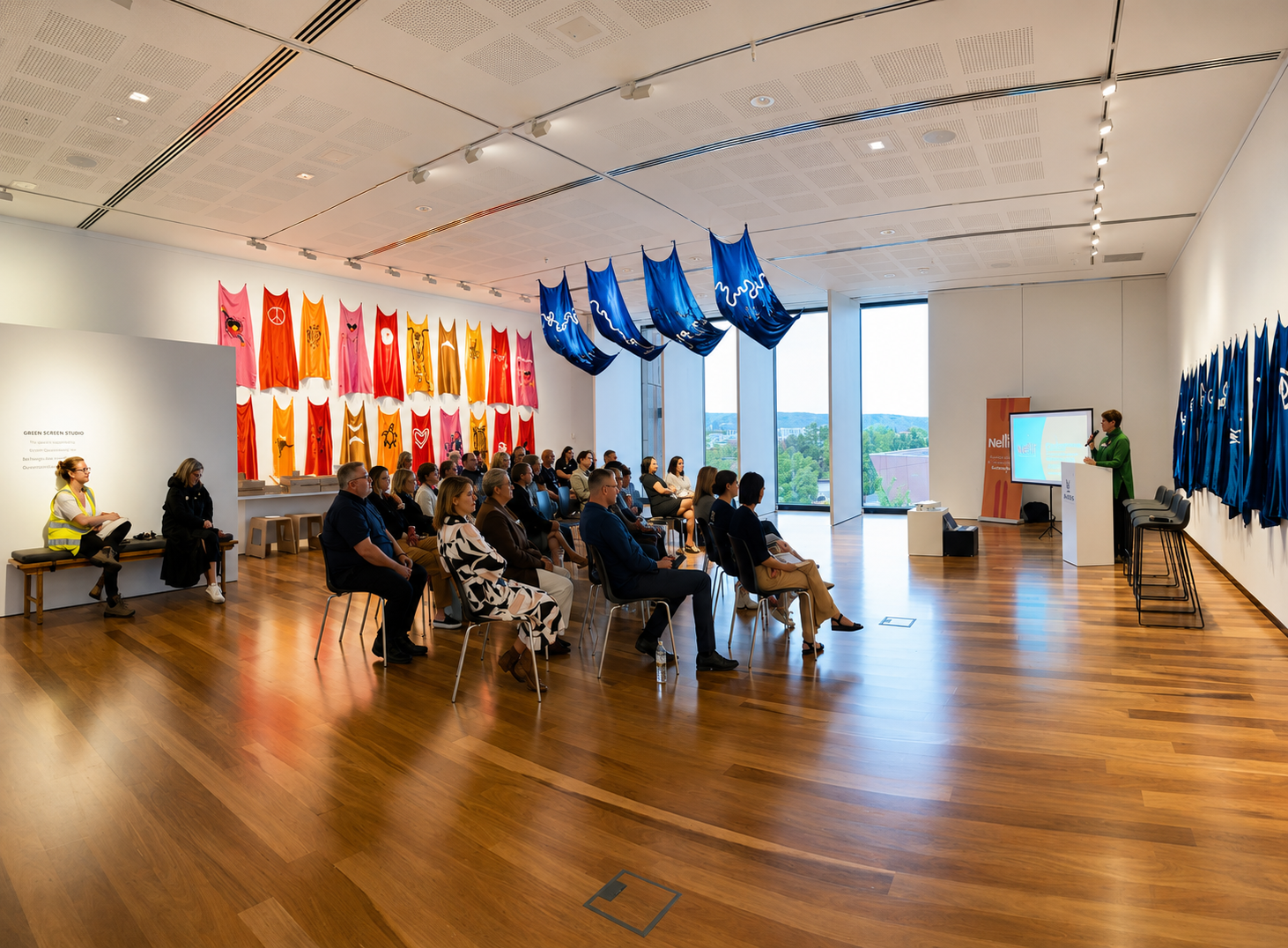 A group of people seated in an art gallery or conference room, listening to a woman giving a presentation at a podium. The room features large windows, colorful banners on the walls, and blue fabric hanging from the ceiling.