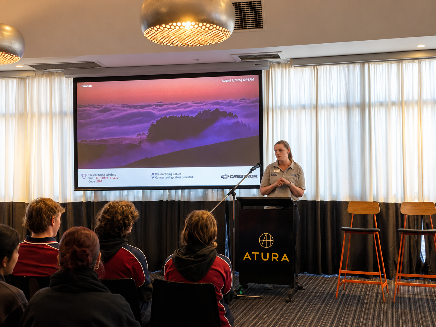 A woman giving a presentation to an audience in a conference room. The presentation slide shows a landscape with mountains and clouds at sunrise or sunset. The room has white curtains, round ceiling lights, and chairs arranged in rows.