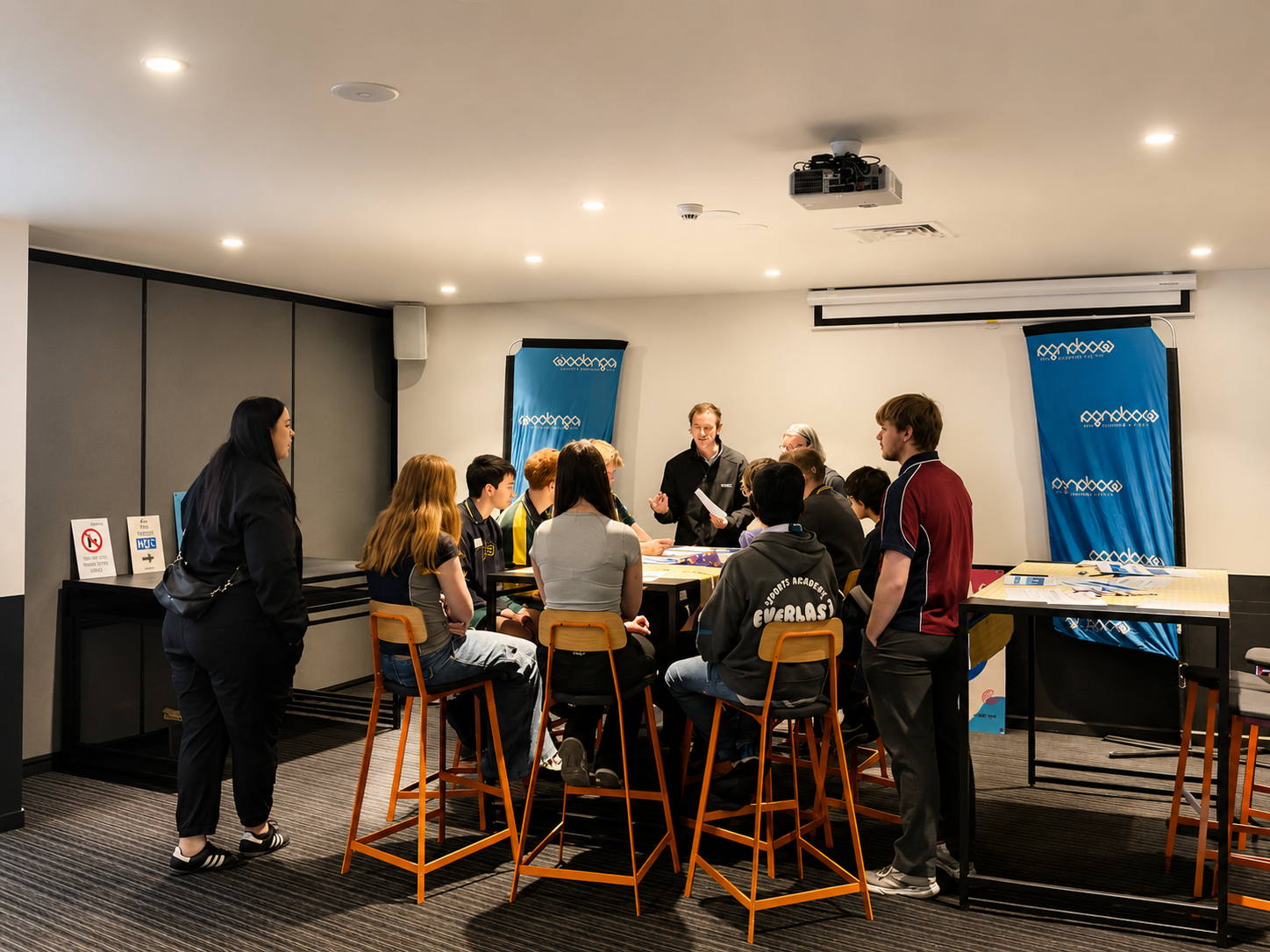 A group of young people sitting around a table in a classroom or seminar room, listening to a man standing and speaking, with blue banners, a projector, and signage visible in the background.