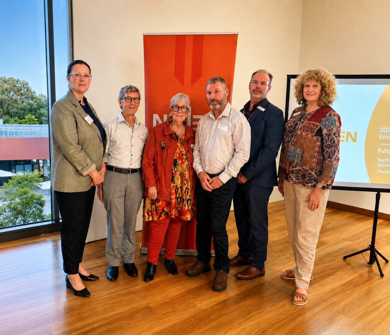Seven people standing together indoors during a presentation or event, with a large window and a presentation screen in the background.