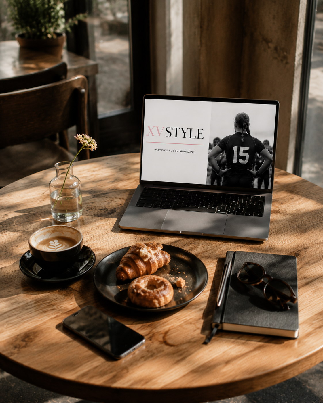 A wooden table in a cafe with a laptop showing a women's rugby magazine, a cup of coffee with latte art, a plate with croissants and a donut, a smartphone, a notebook with sunglasses and a pen, a vase with a flower, near a window with sunlight.