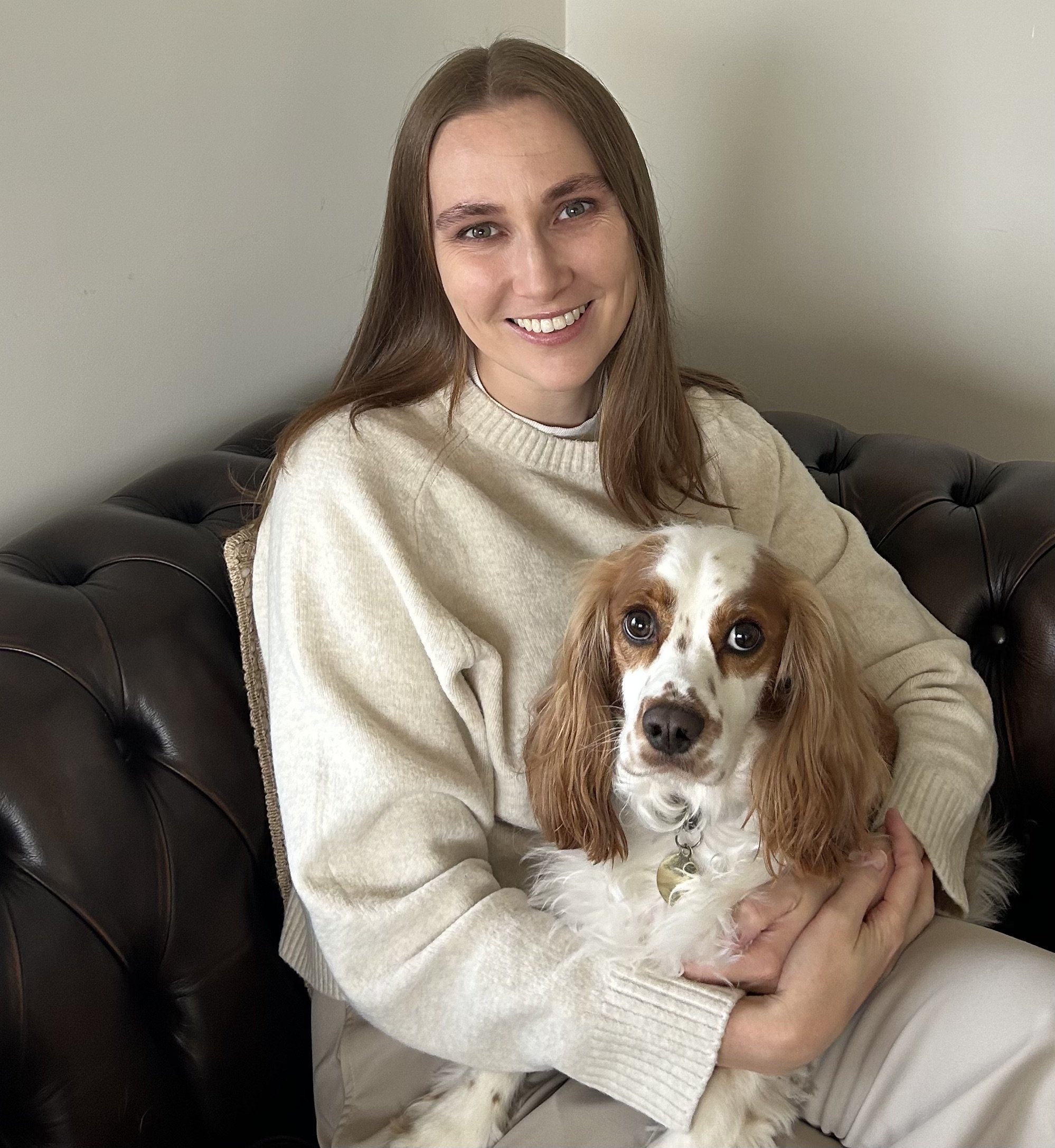 A woman with long brown hair, wearing a cream-colored sweater, sitting on a black leather couch, smiling, holding a white and brown dog.