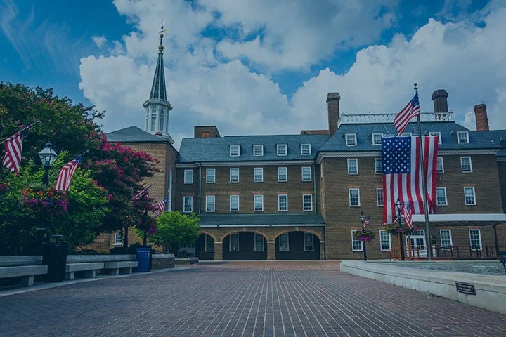 Historic brick building with a tall steeple and several American flags displayed on flagpoles, surrounded by flowering trees and a paved courtyard.