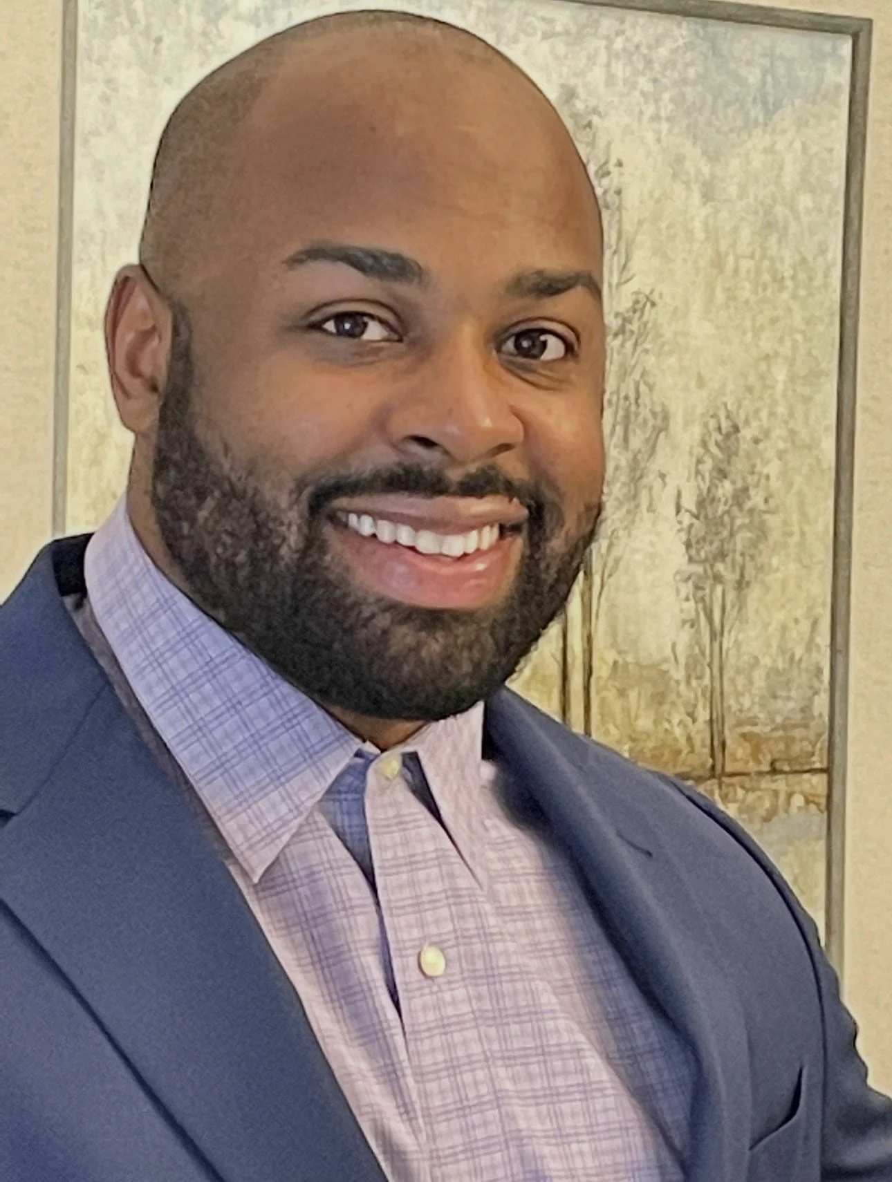 A smiling man with a beard, wearing a blue suit jacket and a pink checkered dress shirt, standing in front of wallpaper with trees.