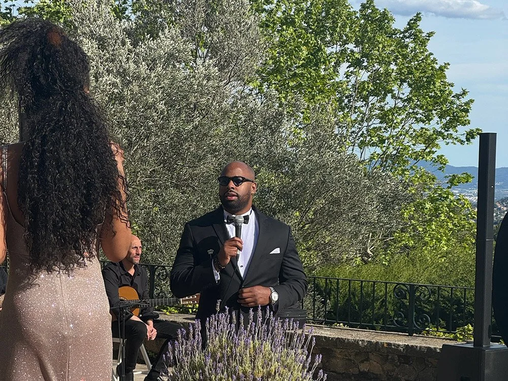 A man in a black tuxedo and sunglasses holding a microphone, speaking outdoors near a woman with long curly hair in a sparkly dress, with a guitarist behind them, greenery, and a scenic view in the background.