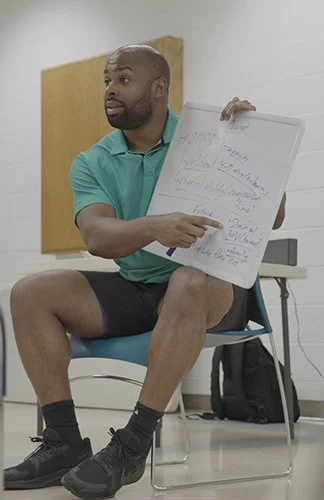 Man sitting on chair holding a whiteboard with handwritten notes, in a room with white walls and a yellow poster or board behind him.