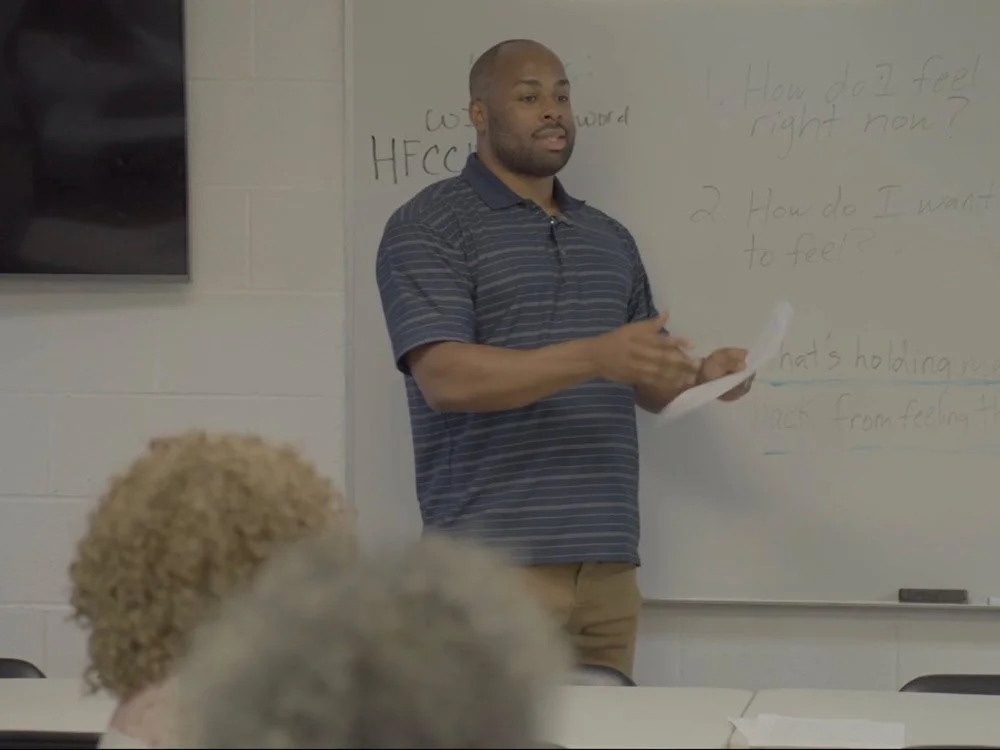Young man teaching in classroom with whiteboard and students, holding papers.