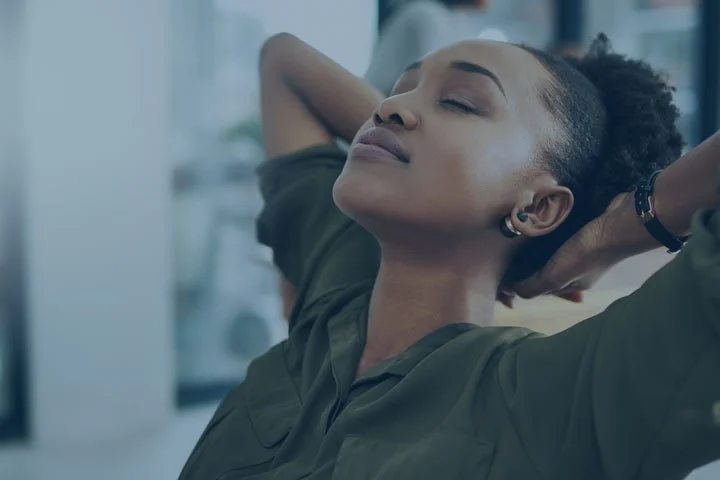 A woman relaxing with her eyes closed and her arms behind her head in an indoor setting.