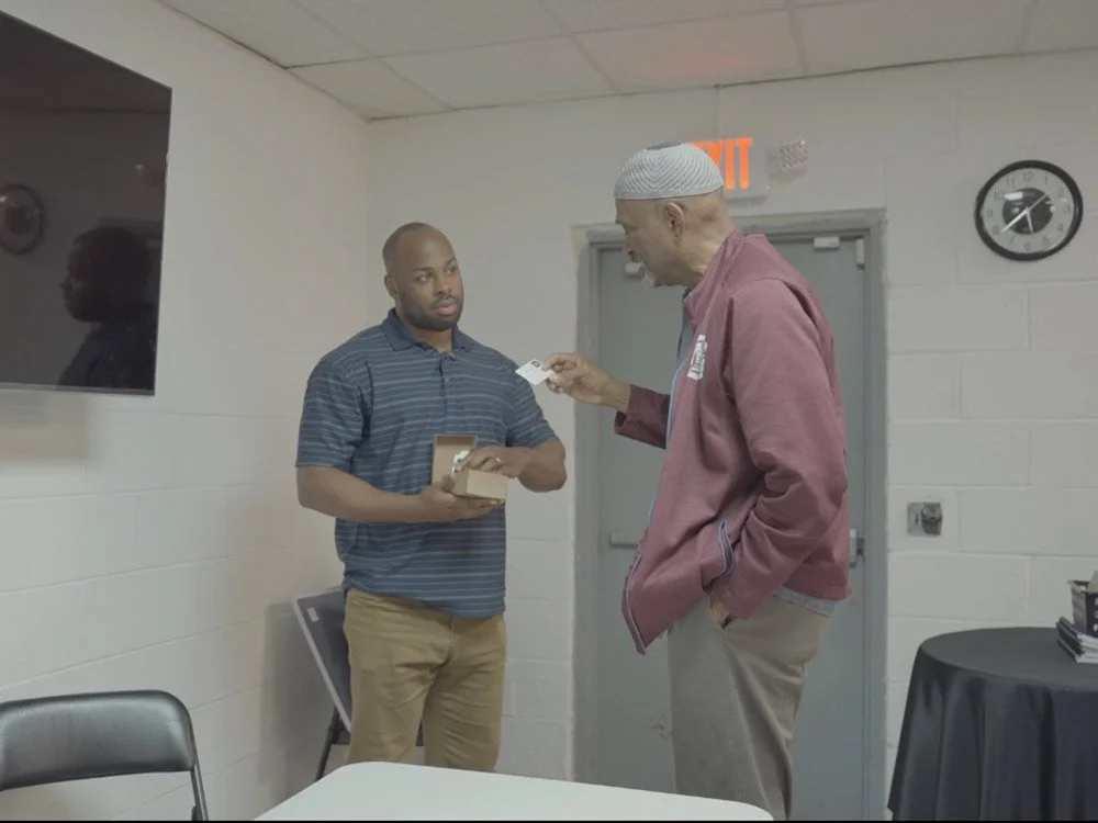 Two men standing in a room, one in a striped polo shirt holding a small box, the other in a maroon jacket and kufi hat handing him a card.