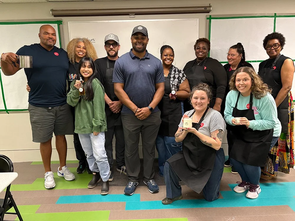 Group of eleven diverse people in a classroom, some holding drinks, posing for a photo. The classroom has a whiteboard and colorful floor tiles, with green tape marking on the walls.
