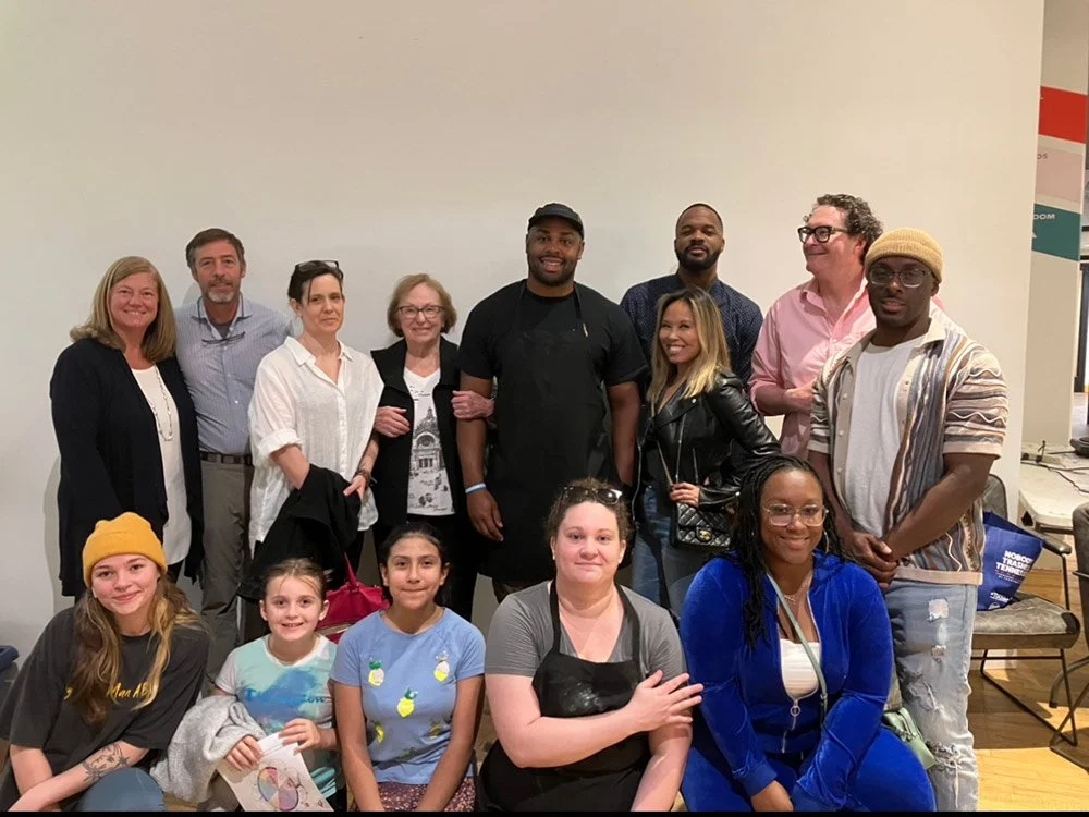 A group of diverse adults and children posing together indoors, some smiling at the camera.