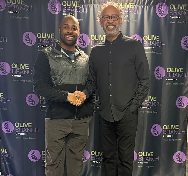 Two men shaking hands in front of a backdrop with the Olive Branch Church logo. One man is wearing a black jacket, and the other is wearing a black shirt and glasses.