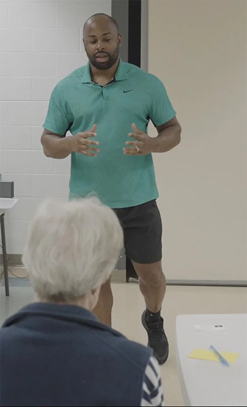 Man in teal polo shirt giving a presentation to an older woman with white hair in a classroom or meeting room.