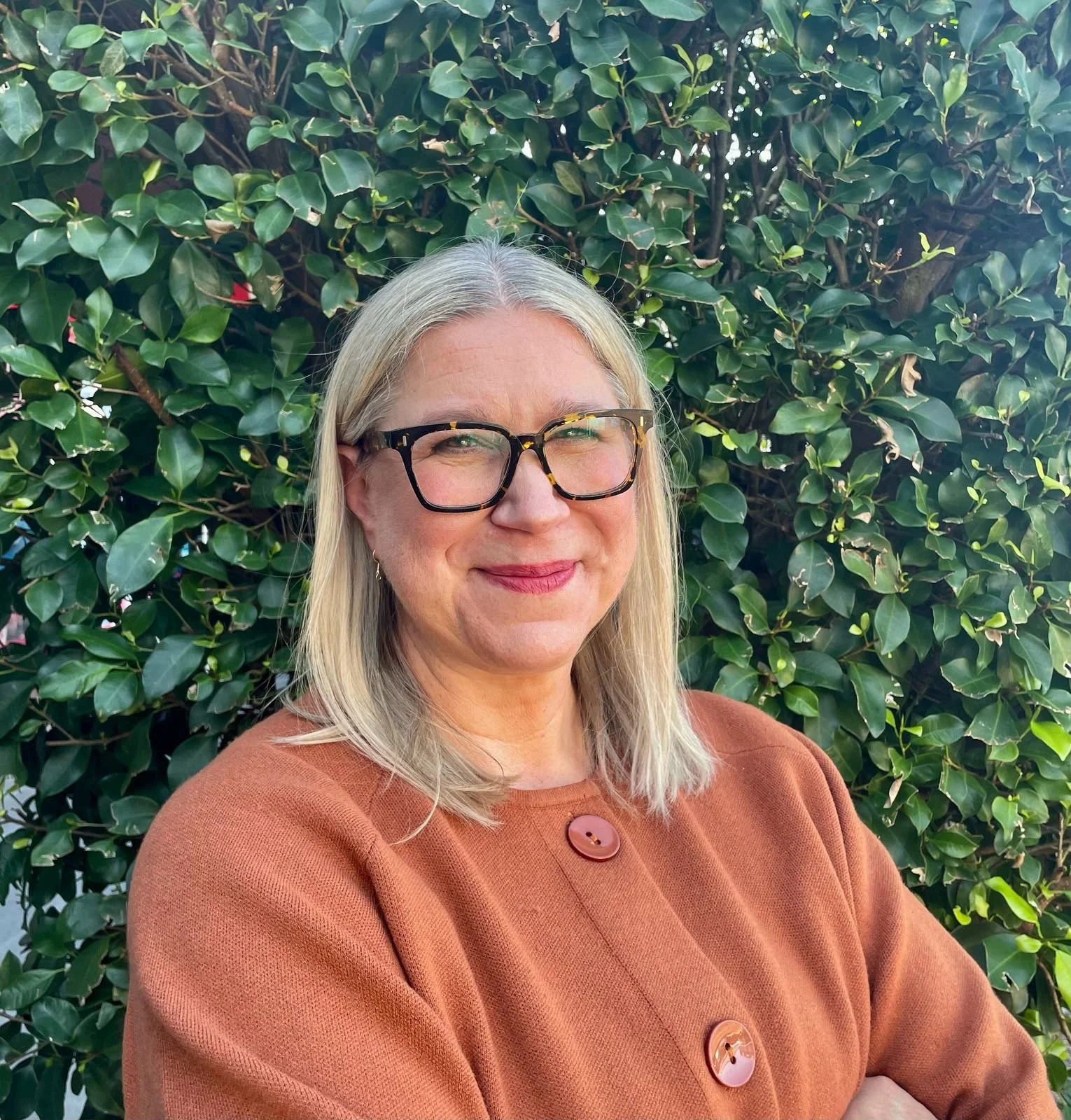 A woman with shoulder-length blonde hair and glasses smiling in front of a large green leafy bush. She is wearing an orange-brown top with large buttons.