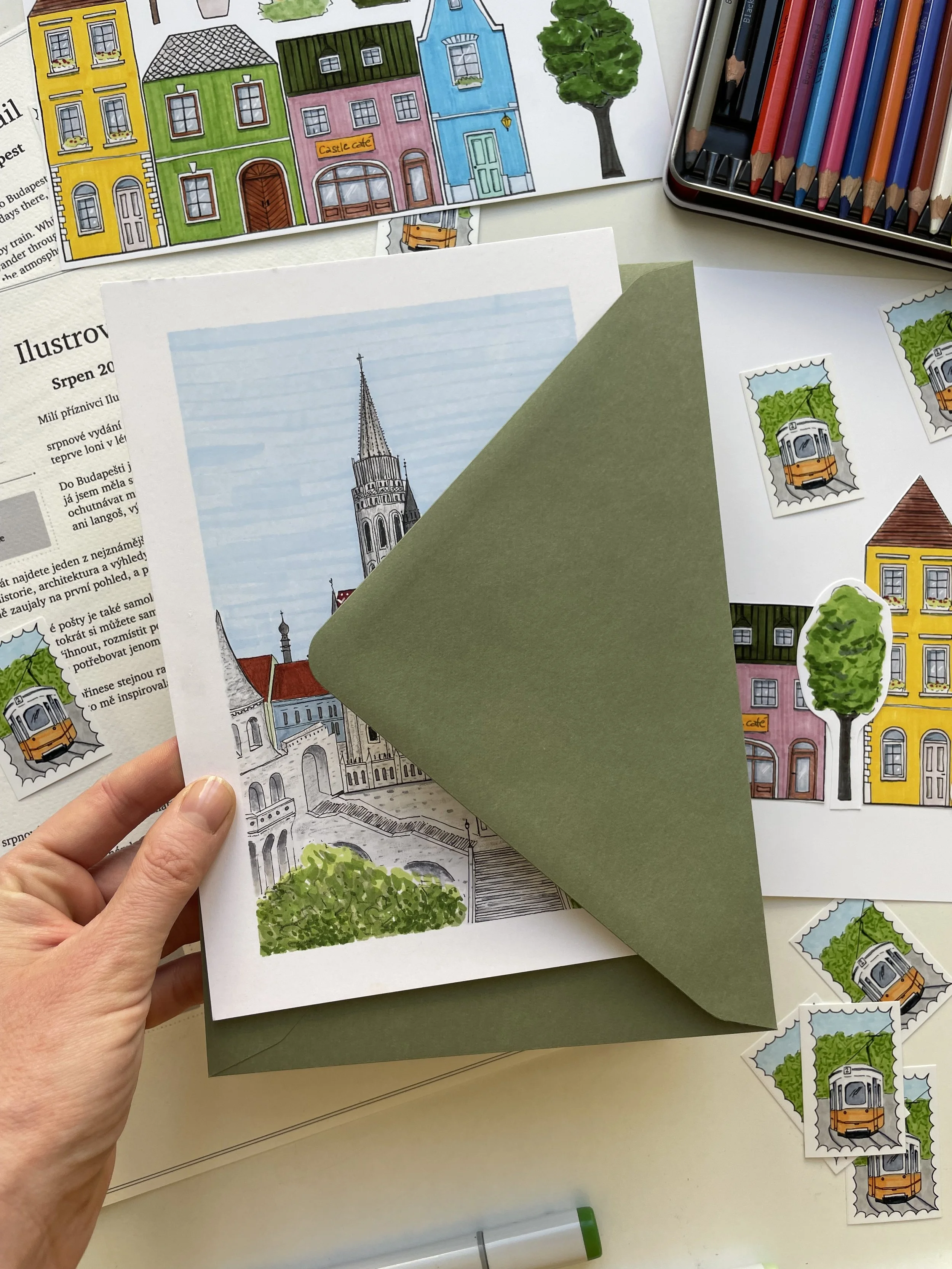A collection of colorful illustrated postcards and stickers featuring houses, trees, a church, and a tram, with a hand holding one postcard and a set of colored pencils visible on the desk.