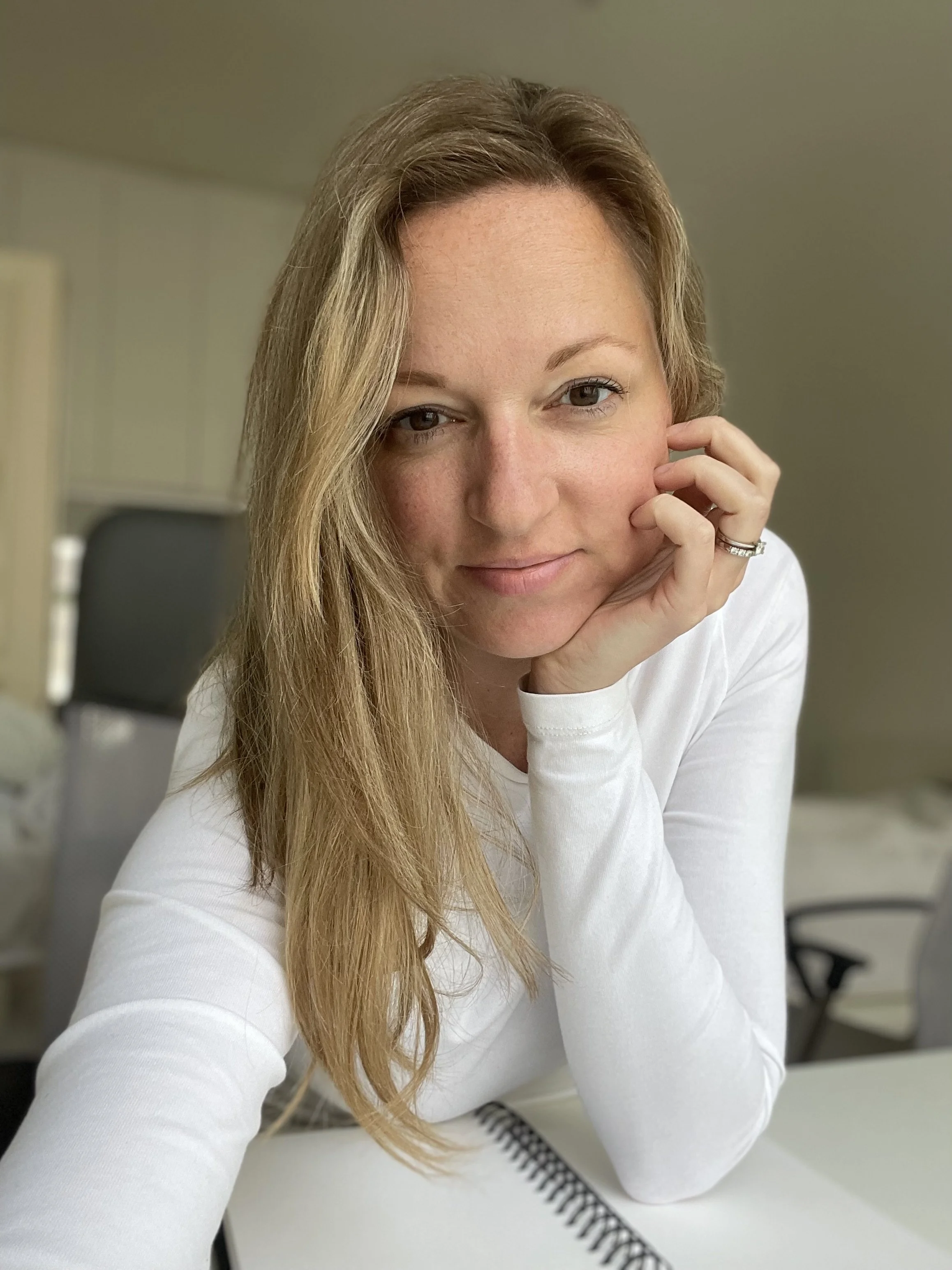 A woman with long blonde hair, wearing a white long-sleeve shirt, resting her head on her hand and smiling softly, sitting at a desk with a spiral notebook and a grey chair in the background.