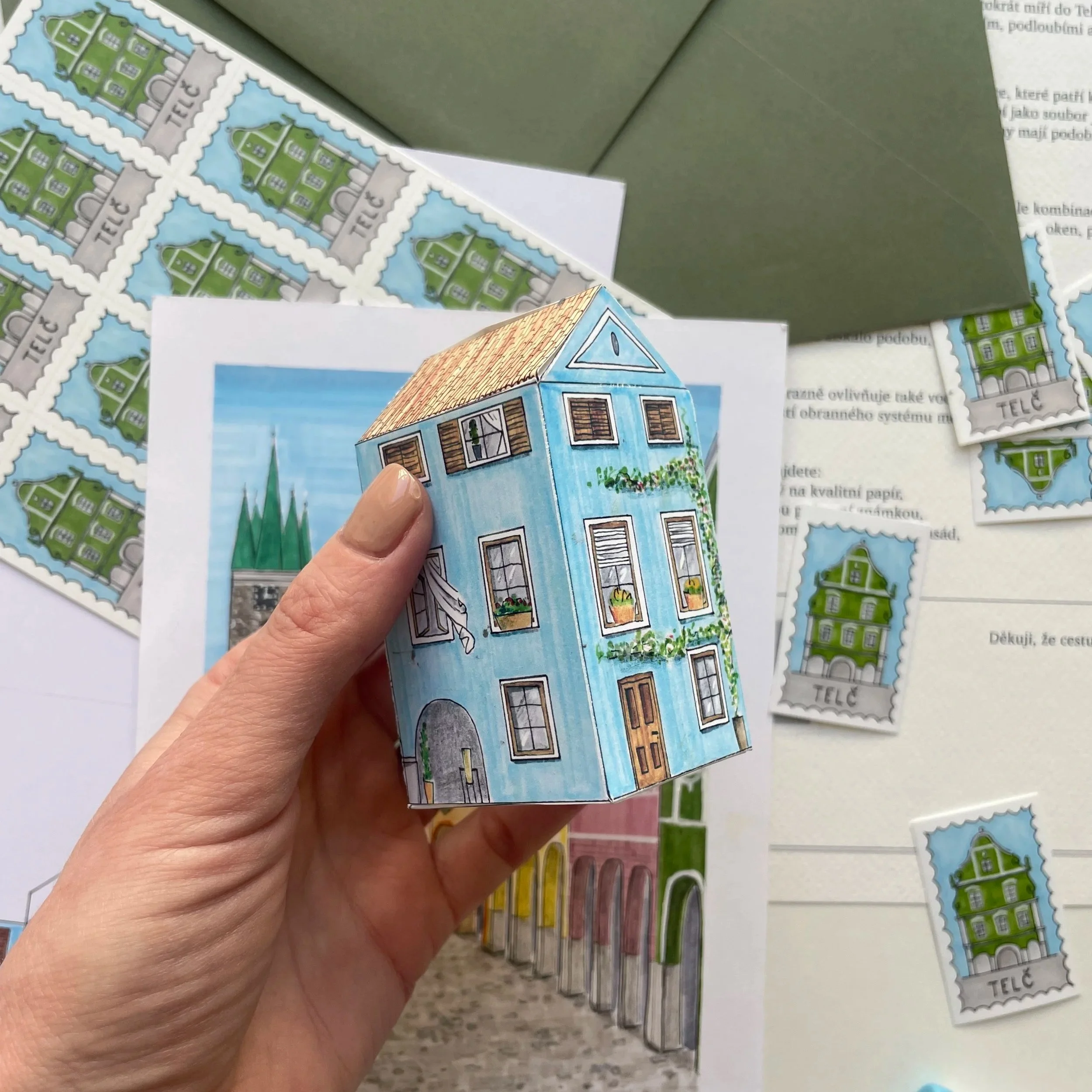 Close-up of a person's hand holding a colorful illustration of a blue house with a red-tiled roof, surrounded by green plants and flowers, with a background of multiple blank postage stamps and printed text.