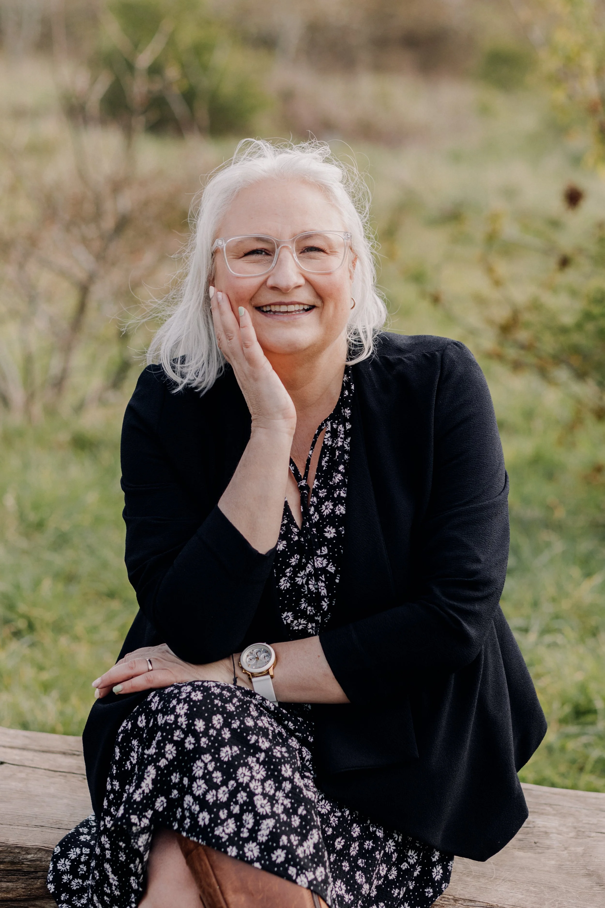 Smiling woman with glasses, sitting outdoors on a wooden bench, wearing a black blazer and floral dress, surrounded by greenery.