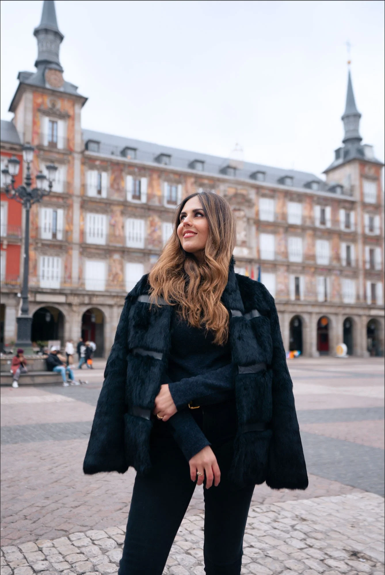 A woman with long wavy hair wearing a black fur coat and black pants standing on a cobblestone square in front of historic European-style buildings with towers and decorative facades, during daytime.