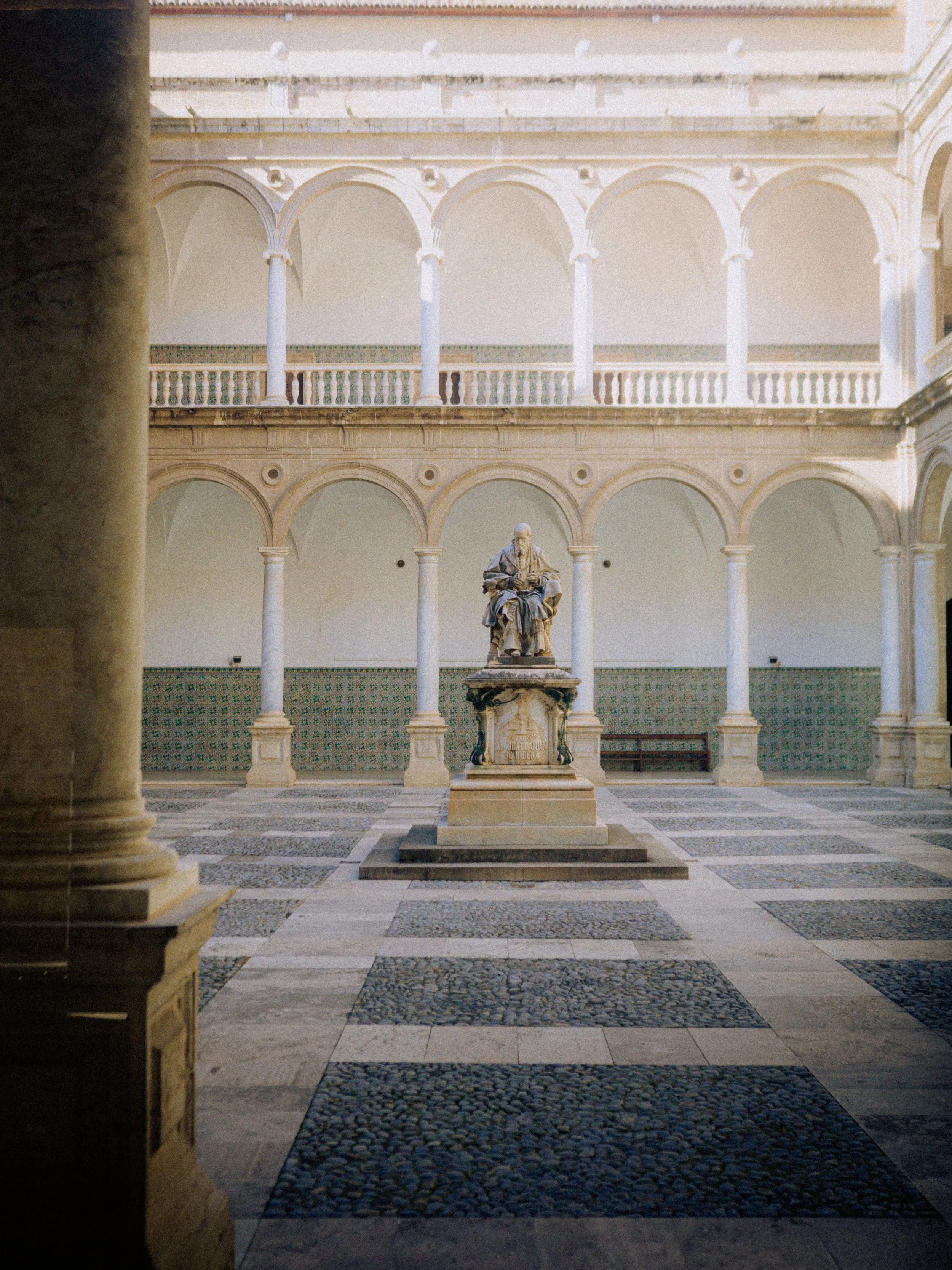 A courtyard with a statue of a seated figure at the center, surrounded by arches and columns in a historic building. The courtyard features a checkered tile floor.
