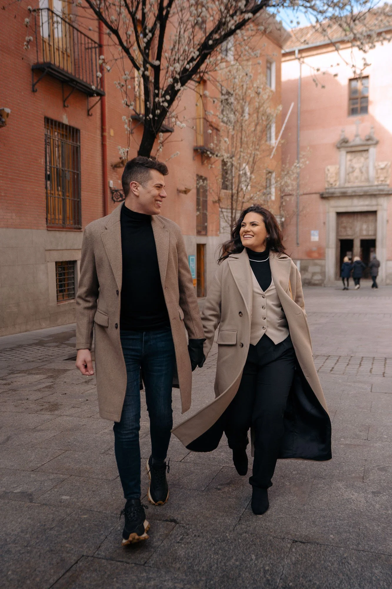 A young man and woman walking hand in hand outdoors in a city square, smiling and looking at each other, surrounded by buildings and blooming trees.