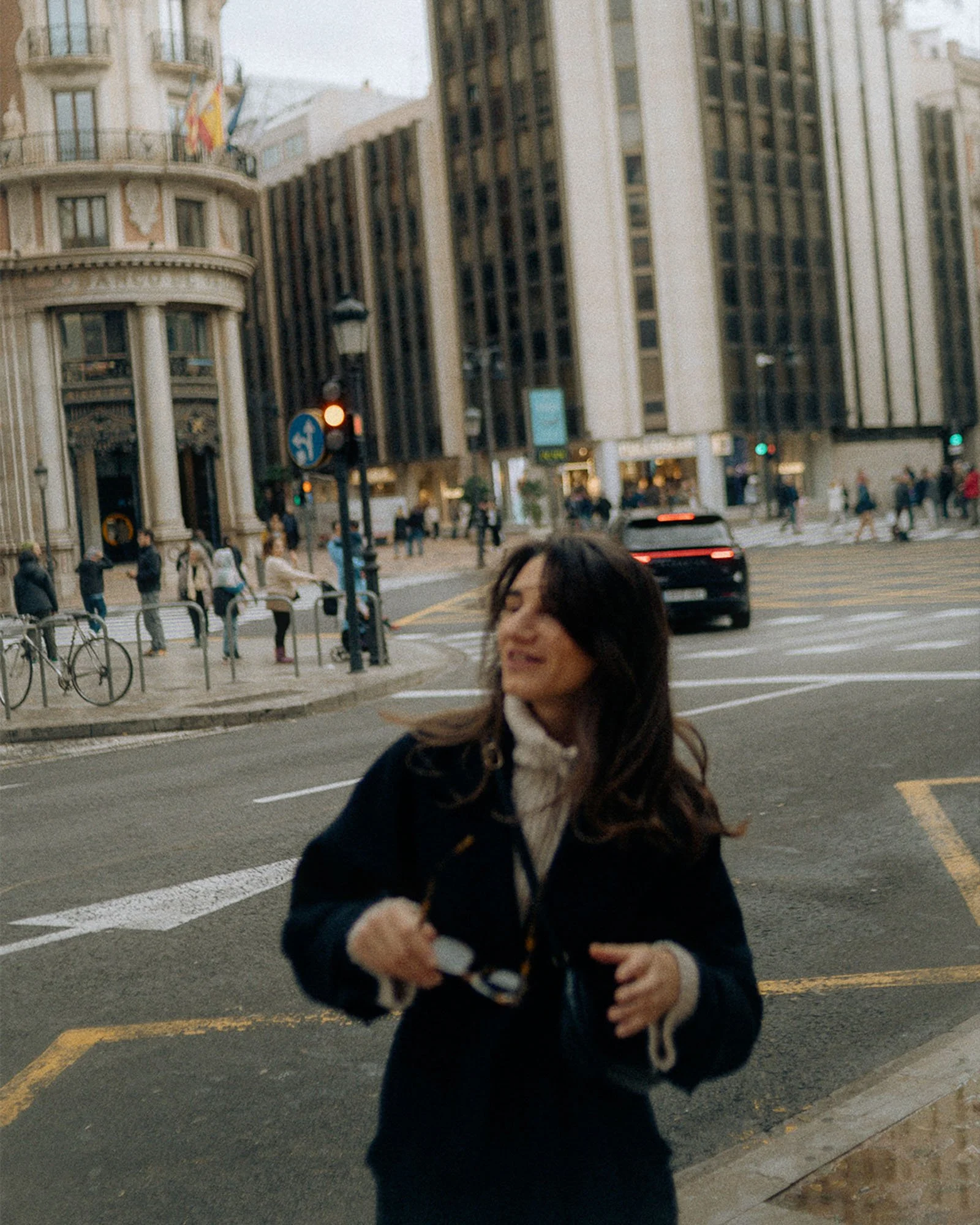 A woman with long dark hair in a black coat is smiling and holding a phone or camera. She is standing on a city street at a crosswalk, with tall buildings and people in the background.