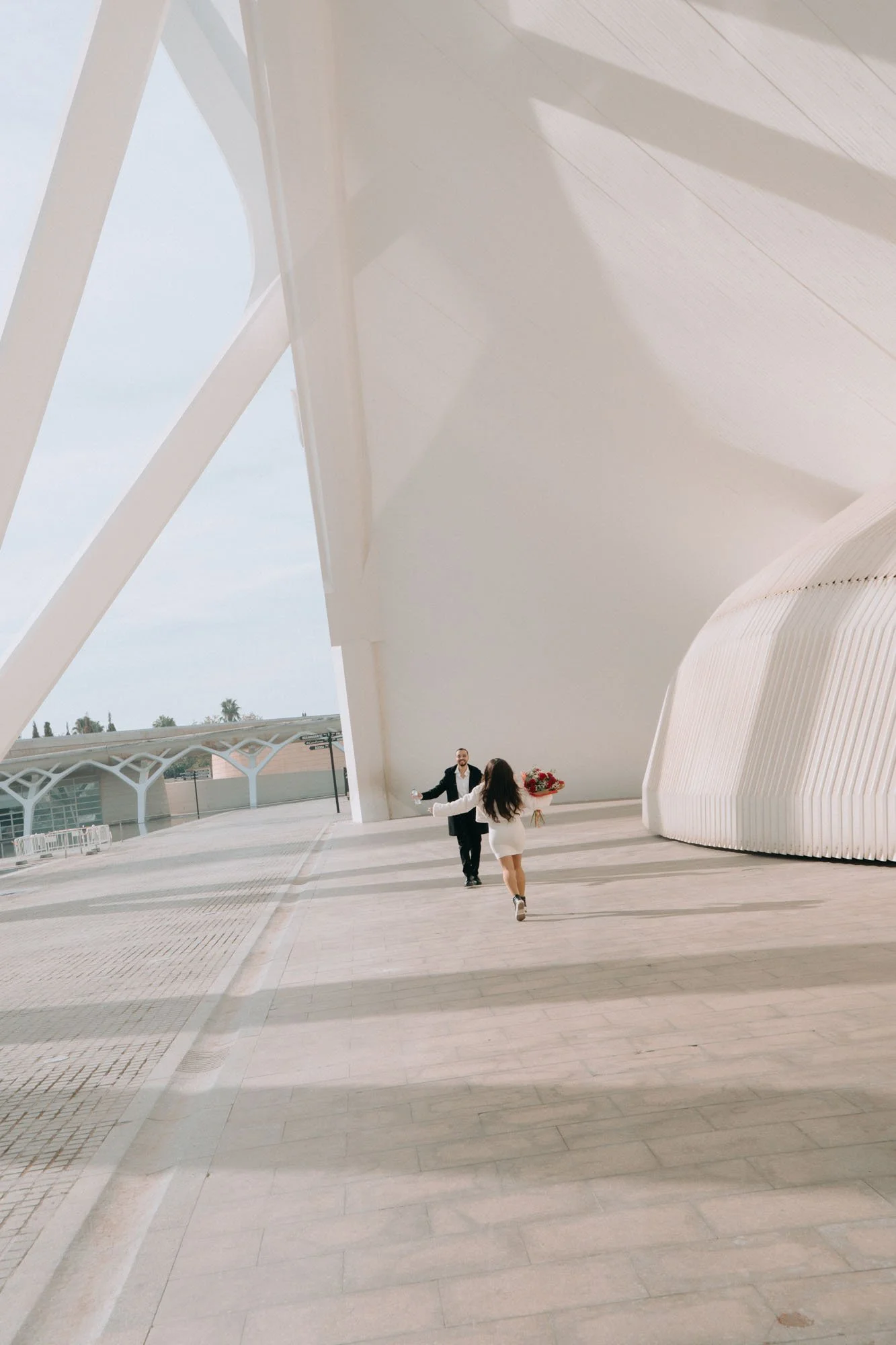 A man and a woman are walking together outside a modern architectural building. The woman is holding a bouquet of flowers, and the man is reaching out towards her. The building features large, white, curved structural elements and a spacious, open ar