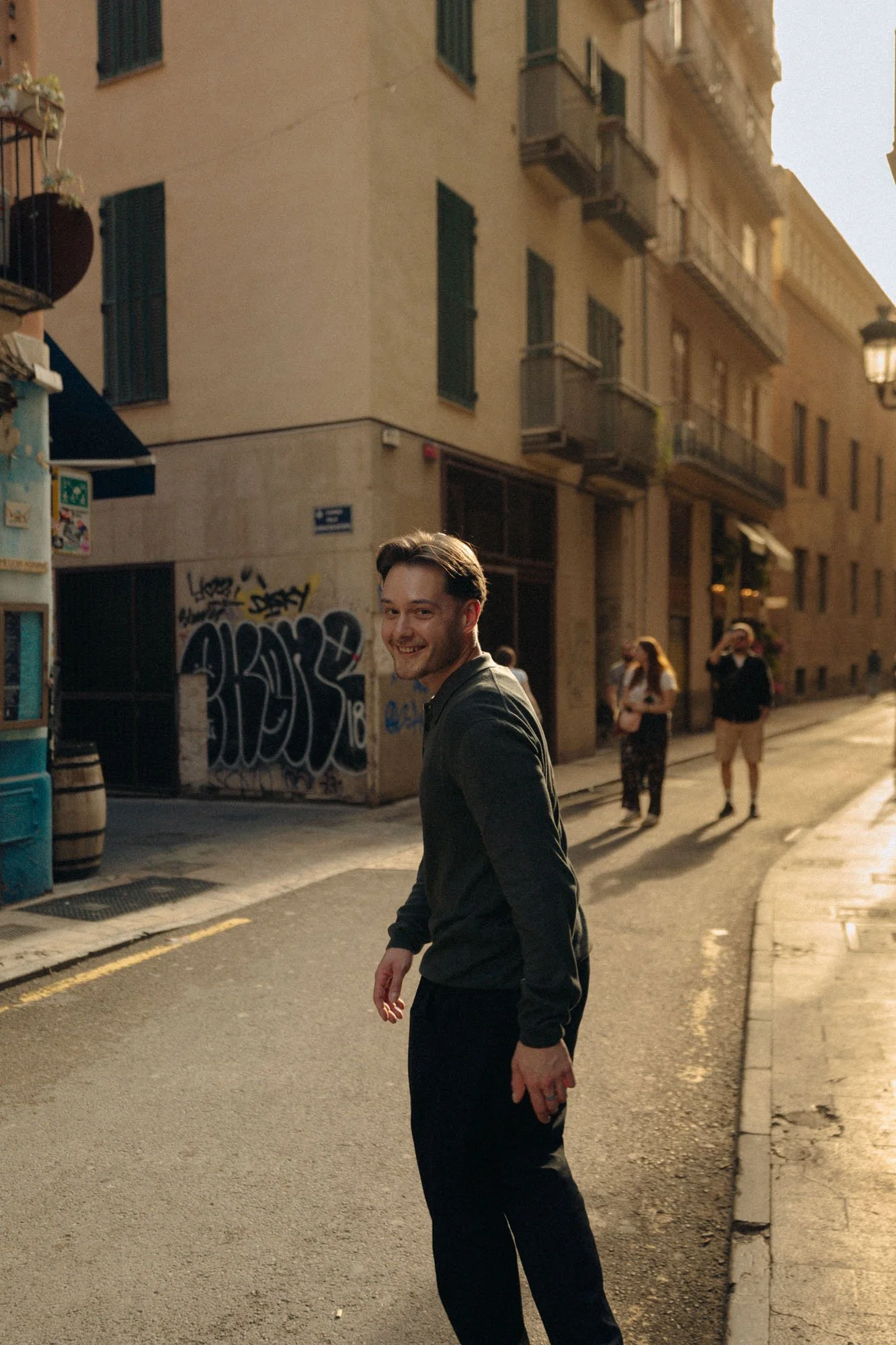 A young man with dark hair and a beard smiling while walking on a city street during the evening or late afternoon. Behind him, there are a few pedestrians and a background of beige buildings with balconies and graffiti.