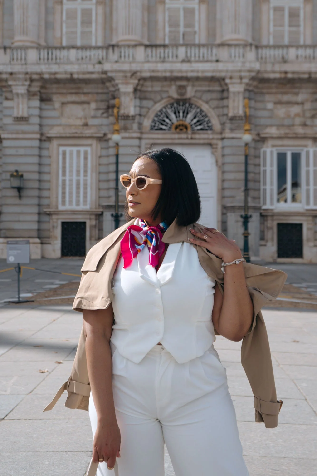 A woman with short black hair wearing beige sunglasses, a beige trench coat draped over her shoulders, a white shirt with a colorful scarf tied around her neck, and white pants, standing outside in front of an historic stone building with ornate wind
