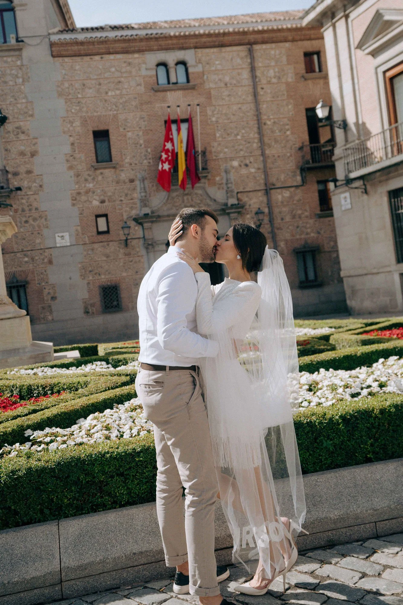 A newlywed couple sharing a kiss outdoors in front of a historic brick building with flags, during daytime. The bride wears a white dress and veil, and the groom wears a light-colored suit with rolled-up sleeves.