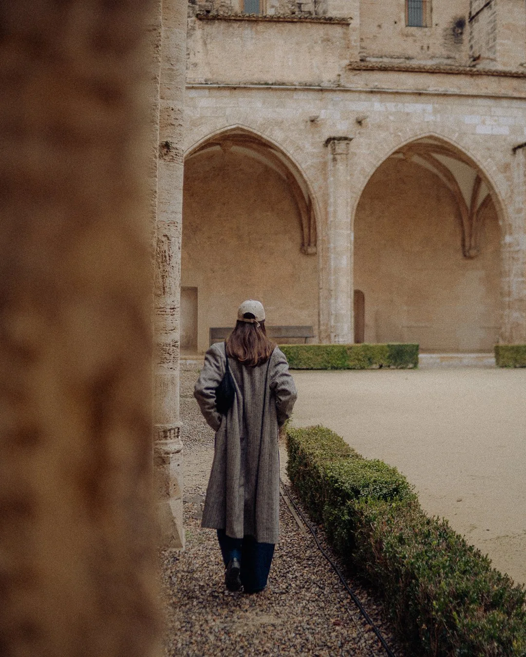 A person with long hair wearing a grey coat, black pants, and a baseball cap is walking through an old stone courtyard with arches and greenery.