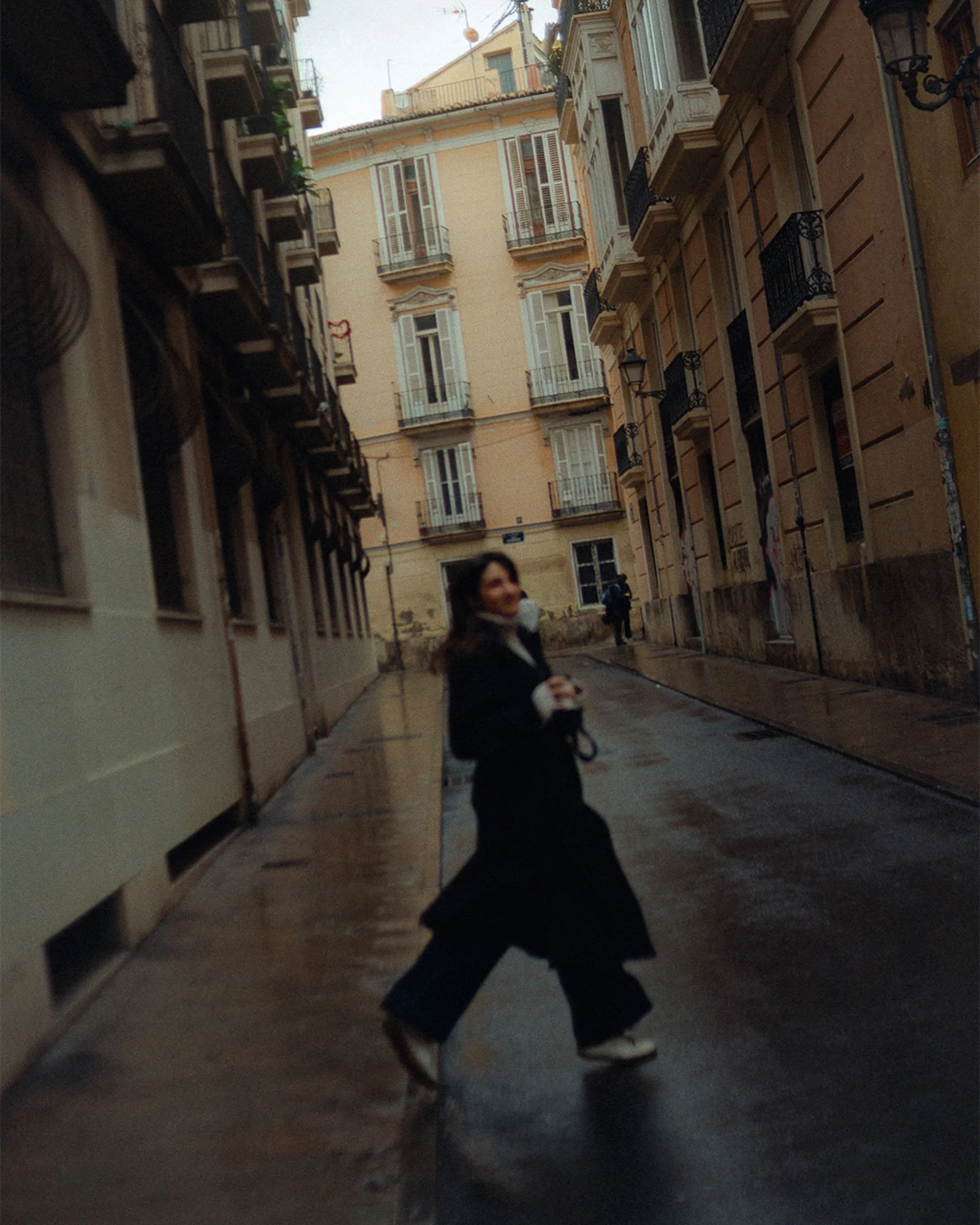 A woman with dark hair, wearing a black coat and white shoes, walking along a narrow street with European-style buildings with balconies and lamps. The street appears wet.