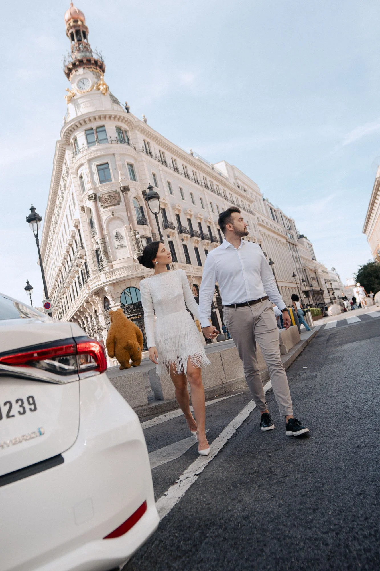 A well-dressed couple walking hand in hand on a city street with historic architecture, street lamps, and a cloudy sky in the background. A dog with fluffy brown fur is walking nearby, and a white car is parked in the foreground.