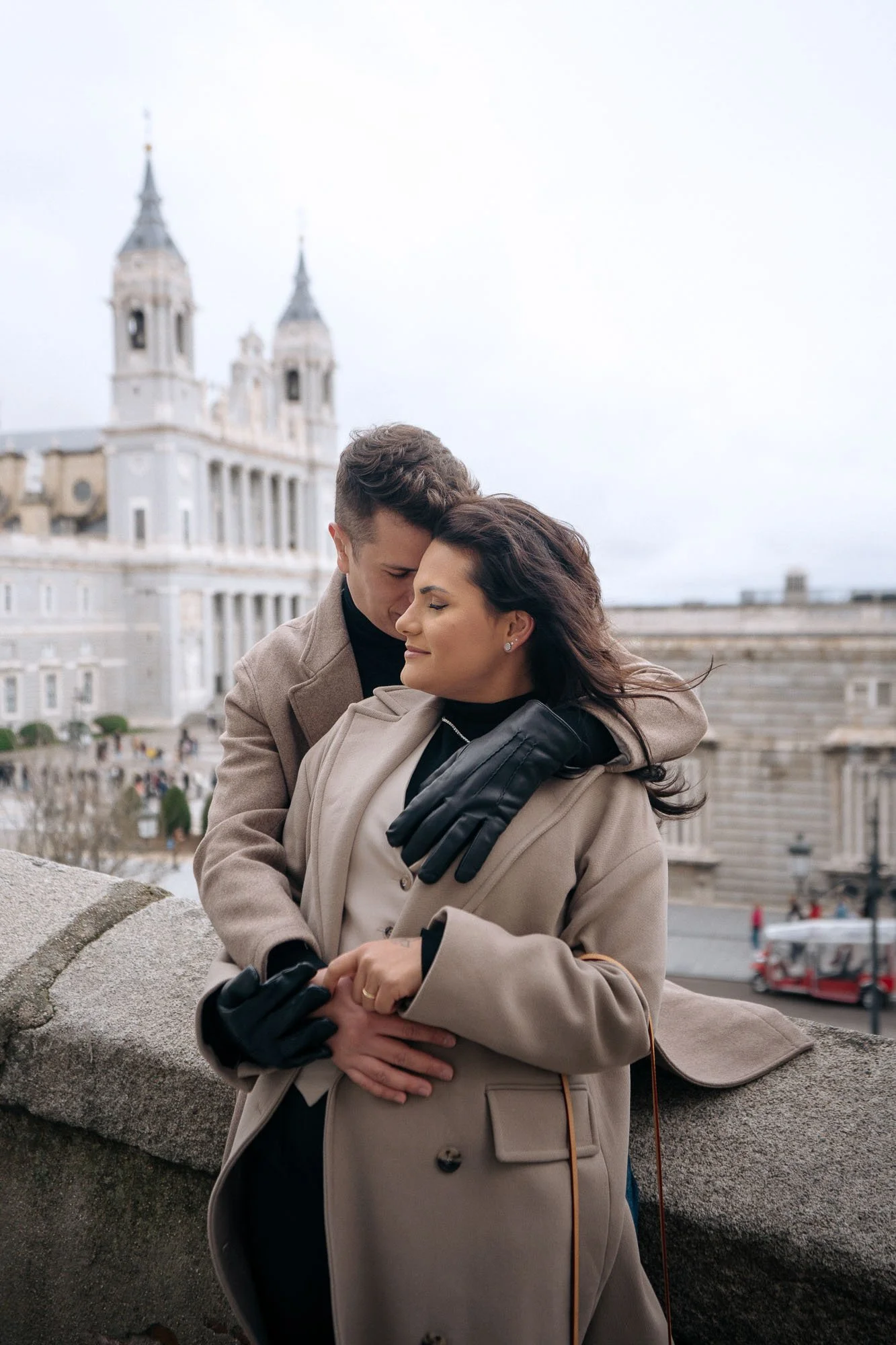 A loving couple sharing a tender moment on a bridge in front of a historic building with twin towers, in a city under an overcast sky.