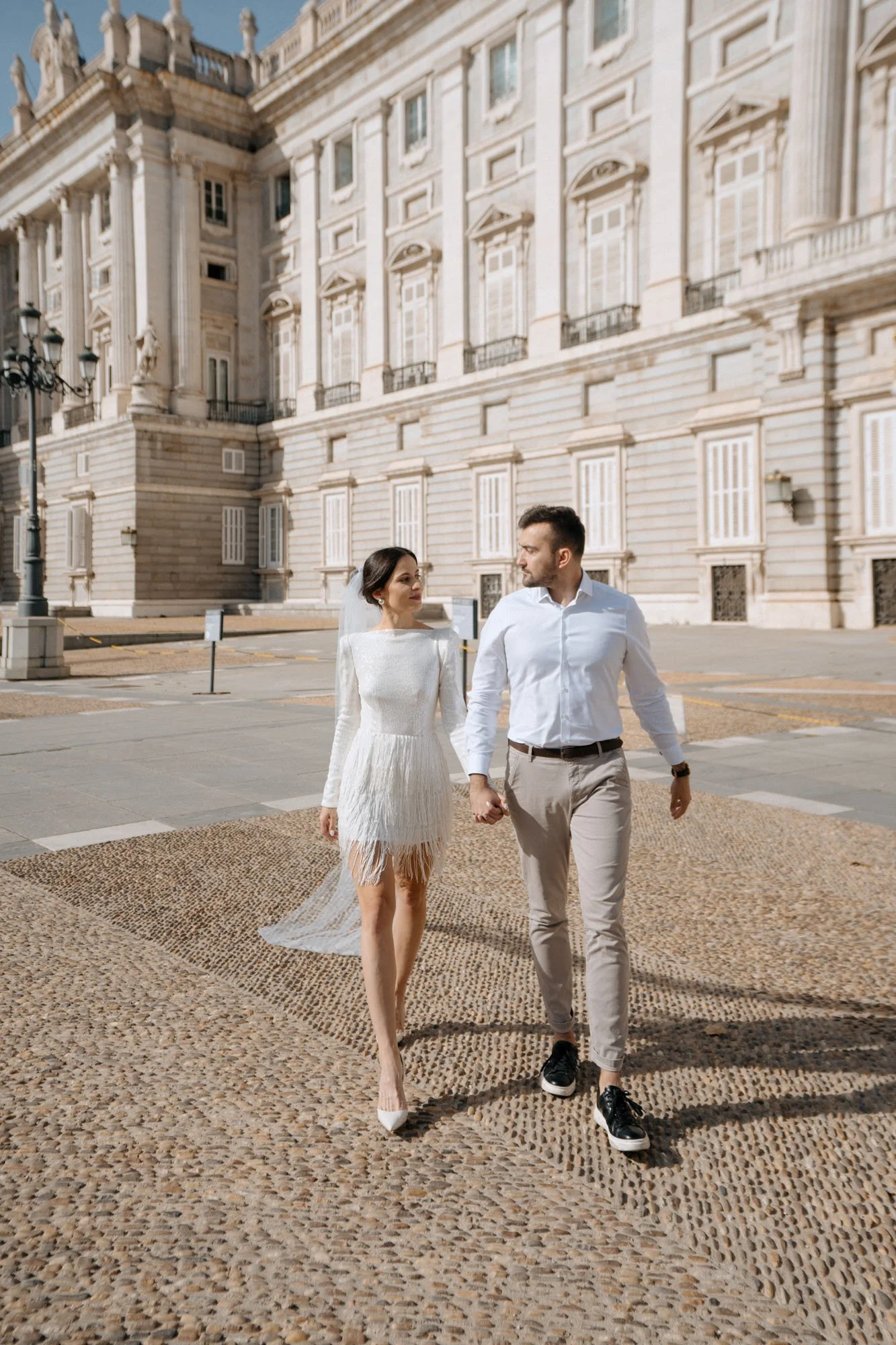 A newlywed couple walking hand in hand in front of a historic building with tall, ornate windows and classical architecture. The bride is wearing a white wedding dress with fringe details, and the groom is dressed in a white shirt and beige pants.