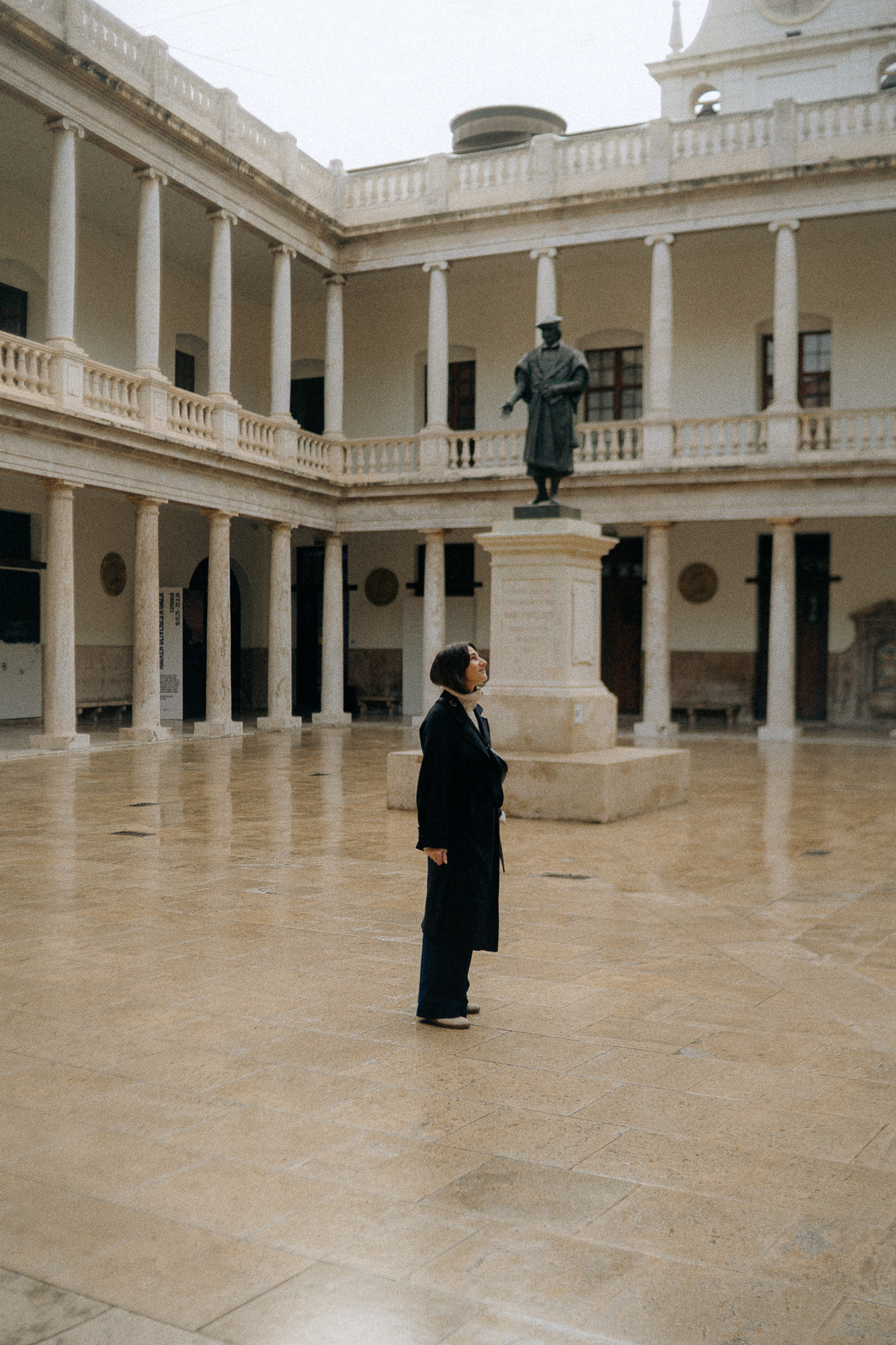 A woman standing inside a large, open courtyard with a two-story colonnade surrounding it. A statue of a man in historical attire is on a pedestal in the center of the courtyard. The woman is looking up at the statue.