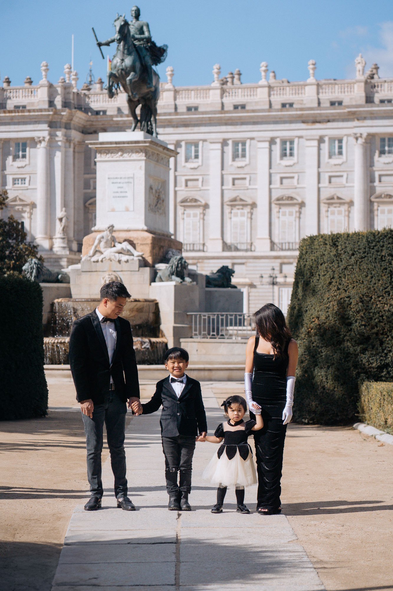 A family of five dressed in formal attire walking hand-in-hand in front of a large white building with a statue of a mounted figure and ornate architectural details.