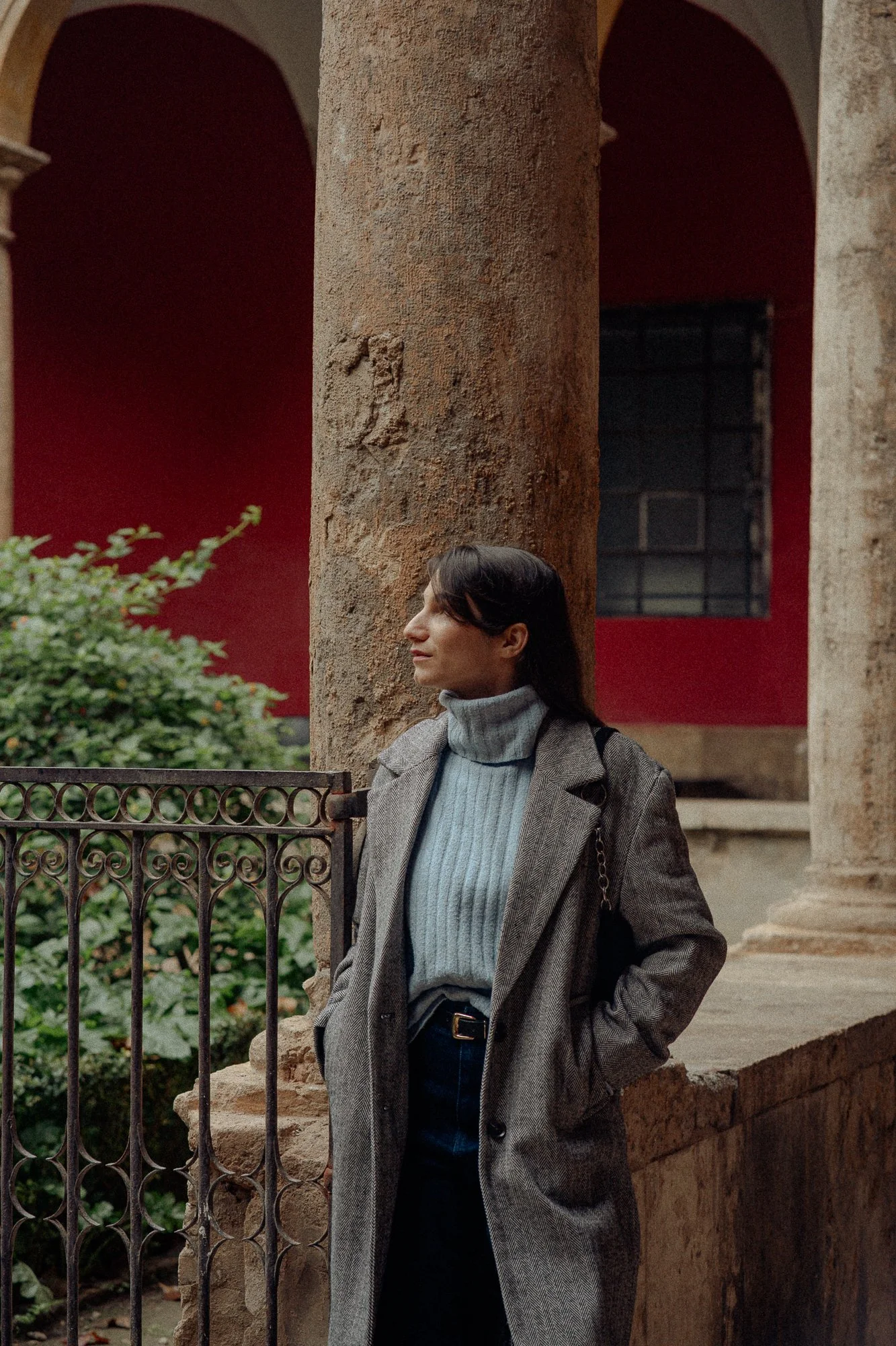 A woman with dark hair wearing a grey turtleneck sweater and a grey checkered coat stands outside near a rusty metal fence and a large stone column surrounded by green foliage and an old red building with arched windows.