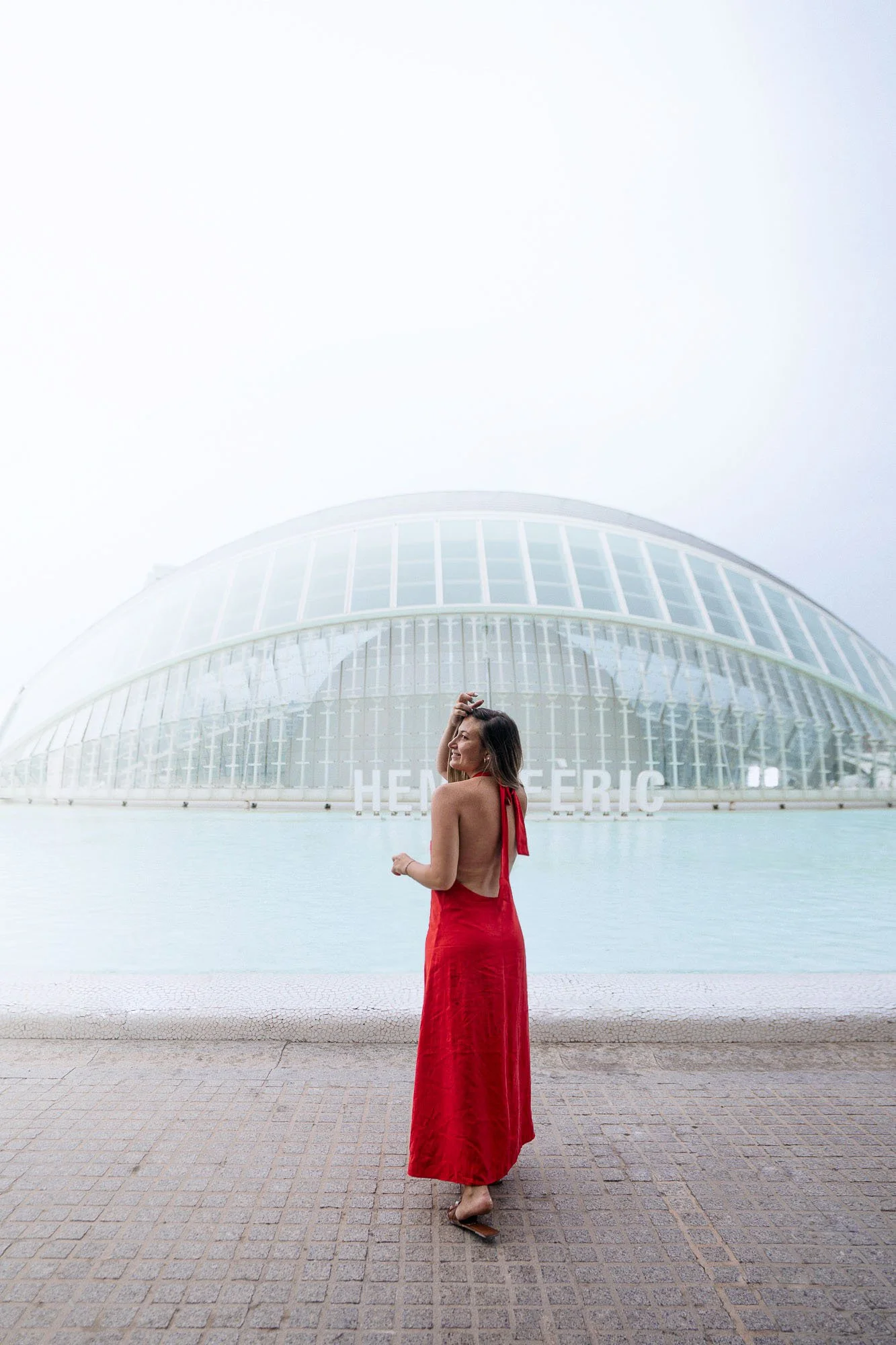 A woman in a red dress is standing by water with her back to the camera, looking over her shoulder, with a modern architectural building behind her that reads 'HENNE FERIC'.
