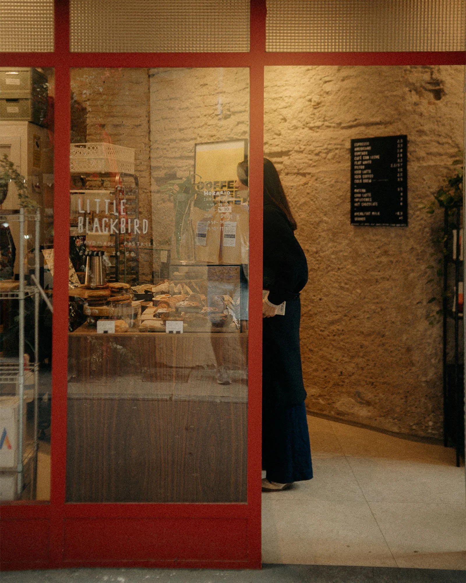 Reflection of a person wearing dark clothing and sneakers inside a bakery in a window with a sign that says "Little Blackbird." Bakery interior with a chalkboard menu and textured wall.